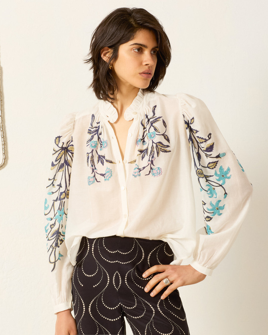 A woman stands against a plain background wearing the Annabel Misted Lily Shirt, featuring a cotton-silk blend with floral embroidery, paired with black pants adorned with a white wavy pattern.