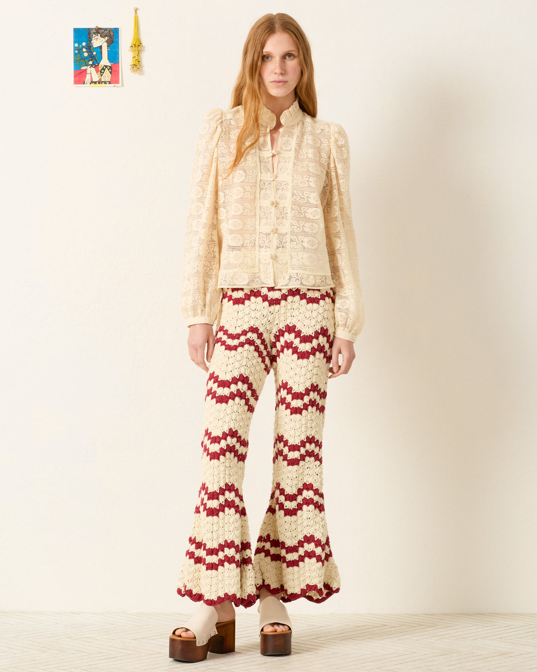 A woman stands against a plain wall, wearing the Artemis Alabaster Lace Shirt, flared crochet pants in cream and red, and brown platform sandals.