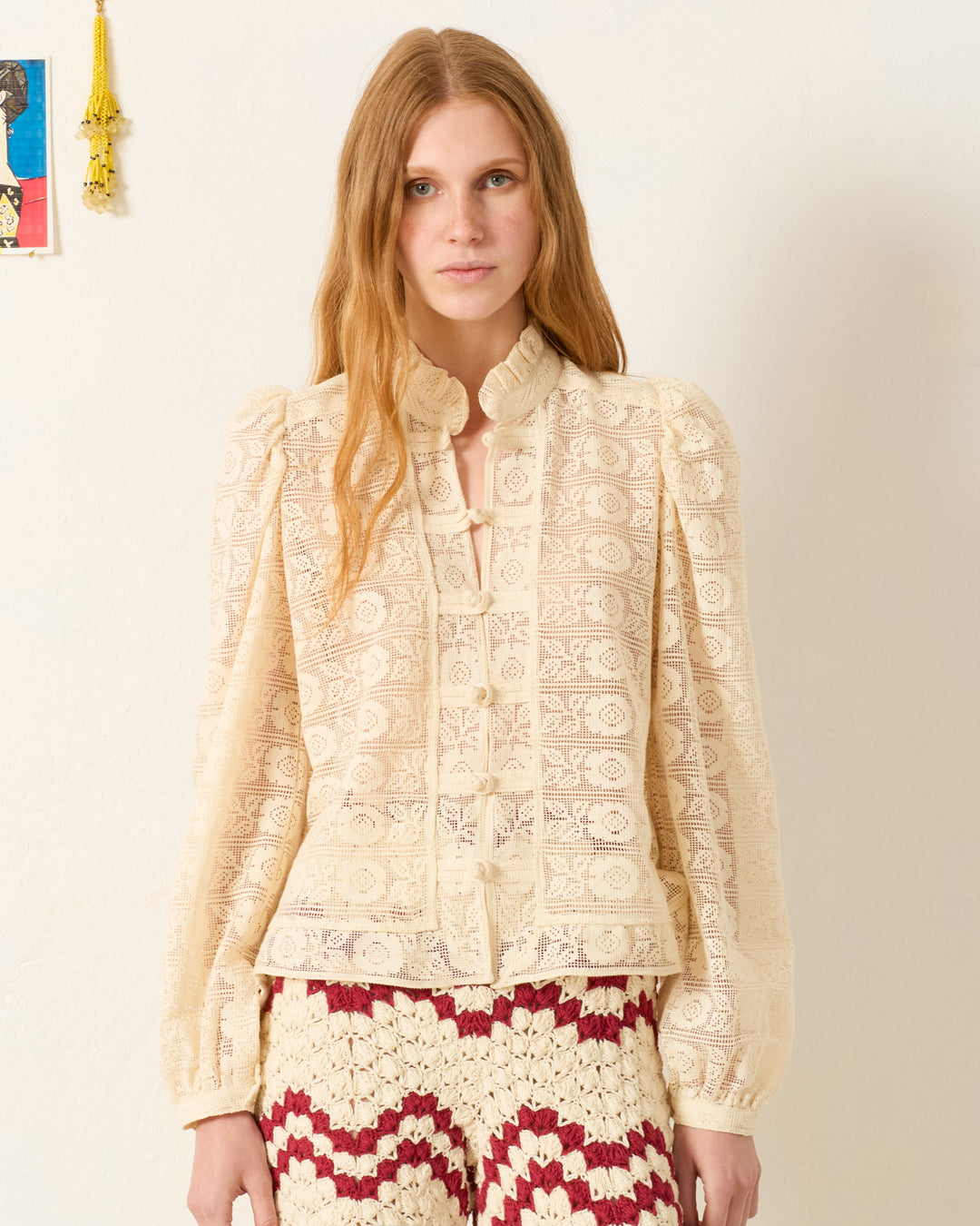 A woman with long, light brown hair wears the Artemis Alabaster Lace Shirt, a cream Victorian-style blouse with floral lace and puffed sleeves, paired with red-and-white crochet pants as she stands against a plain wall.