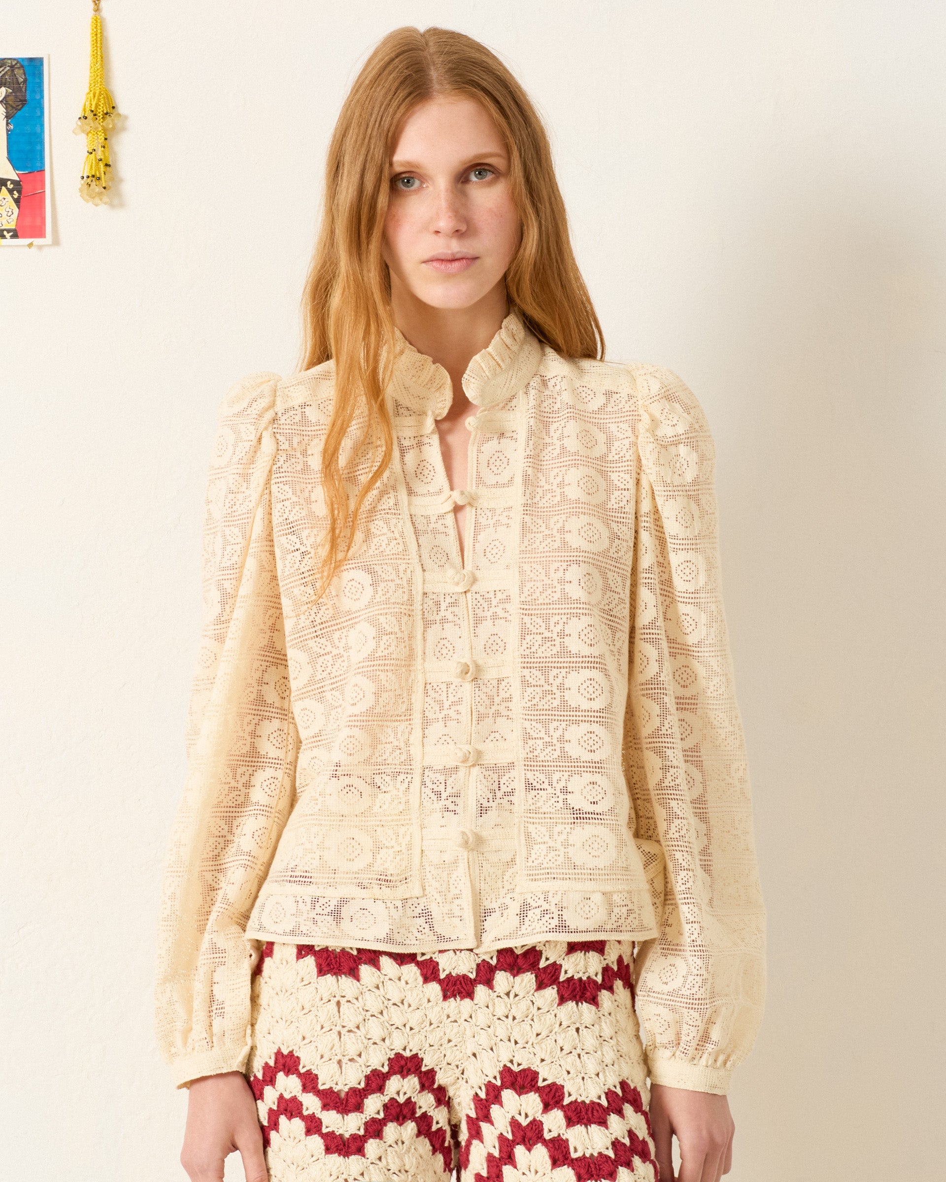 A woman with long, light brown hair wears the Artemis Alabaster Lace Shirt, a cream Victorian-style blouse with floral lace and puffed sleeves, paired with red-and-white crochet pants as she stands against a plain wall.