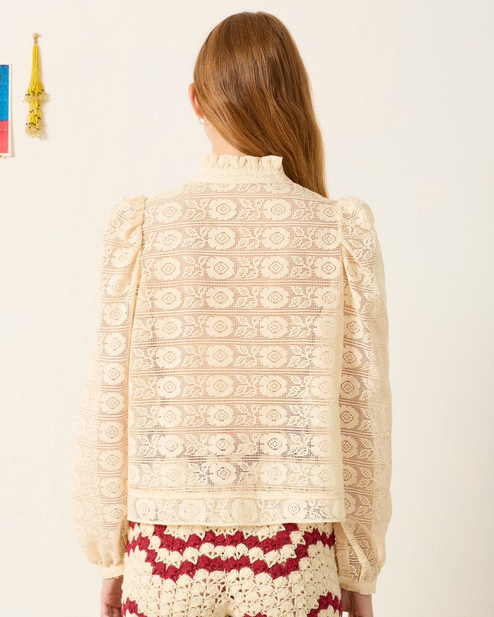 A woman with long brown hair, seen from the back, wears the Artemis Alabaster Lace Shirt with long sleeves and a ruffled collar, paired with a red and cream crocheted skirt.