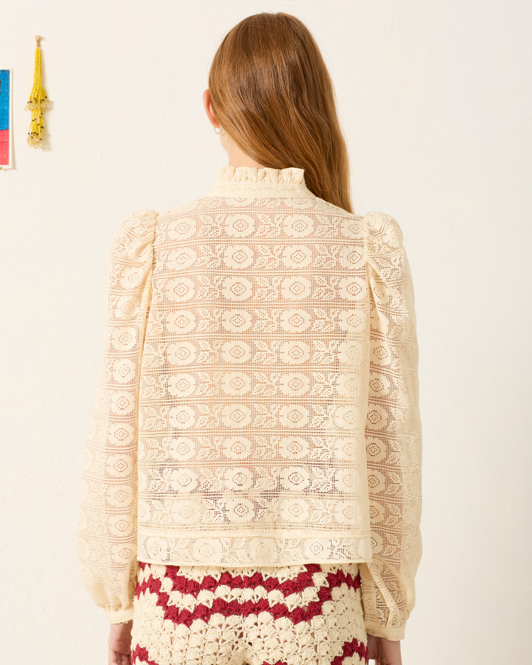 A woman with long brown hair, seen from the back, wears the Artemis Alabaster Lace Shirt with long sleeves and a ruffled collar, paired with a red and cream crocheted skirt.