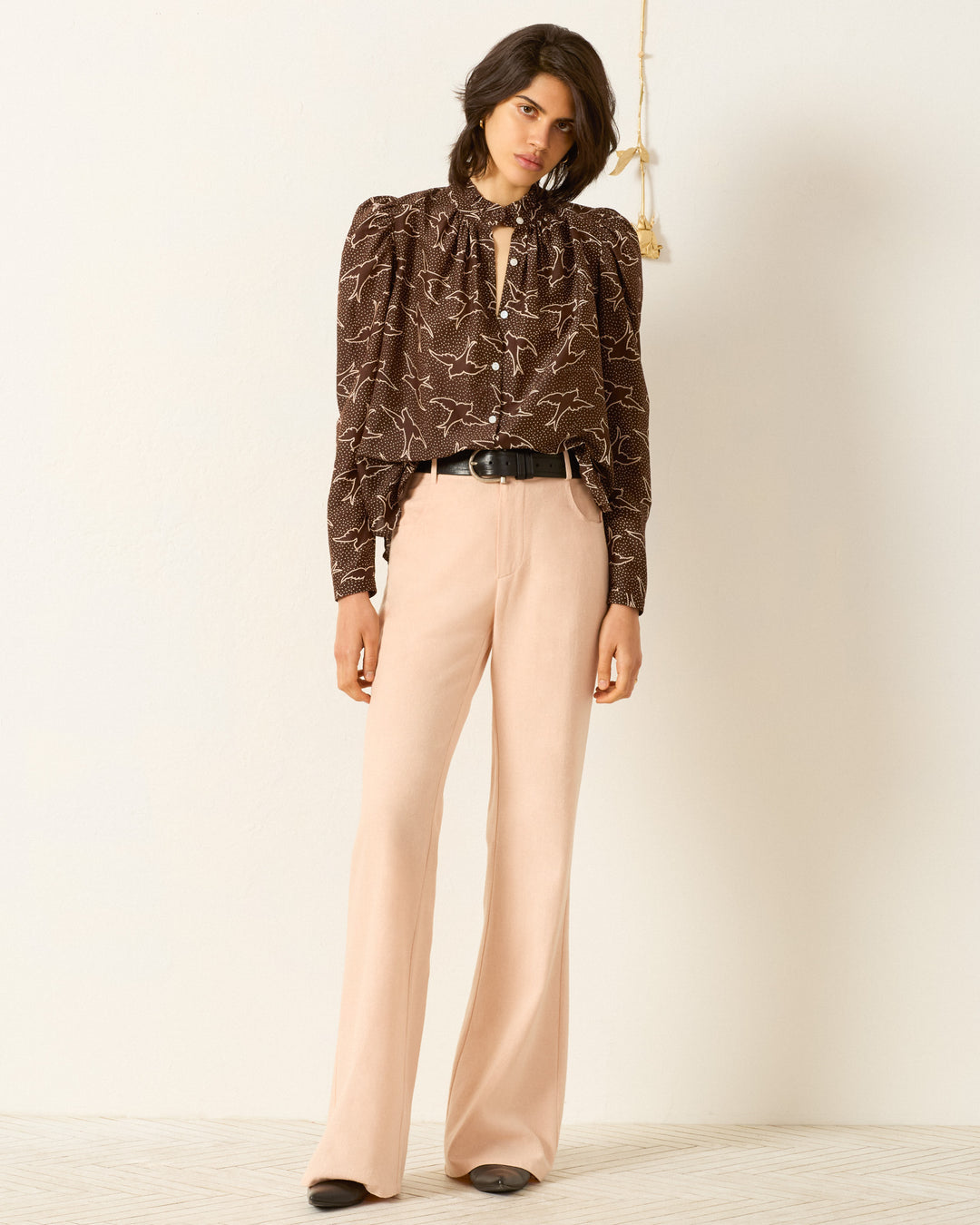 Woman indoors wearing the Artemis Clove Swallow Shirt—a dark brown puff-sleeve blouse with a ruffled collar—styled with beige high-waisted trousers, a black belt, and black-toed shoes.