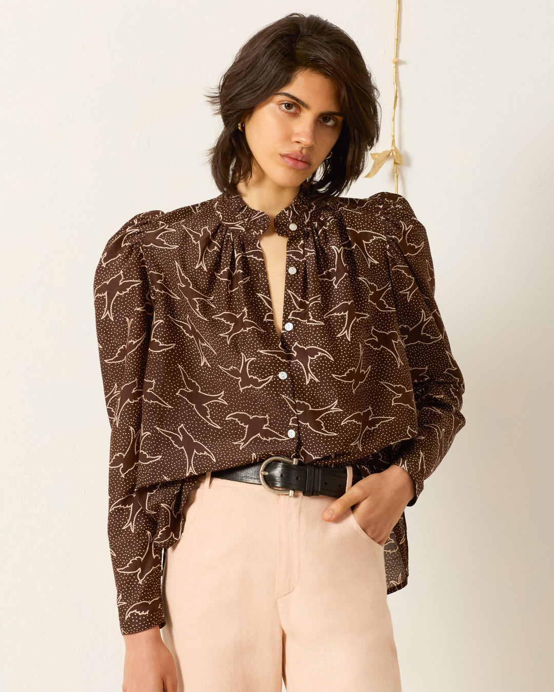 Person wearing the Artemis Clove Swallow Shirt, a brown patterned blouse with puff sleeves and a ruffled collar, paired with light beige pants, standing against a plain light-colored wall.