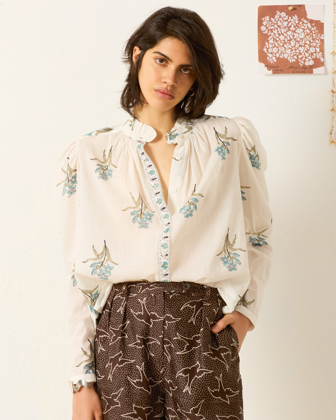 A person stands against a light wall, wearing the Artemis Sky Mist Flower Shirt—a white cotton voile top with a floral motif and puffed sleeves—paired with patterned brown trousers. A small artwork hangs on the wall behind them.