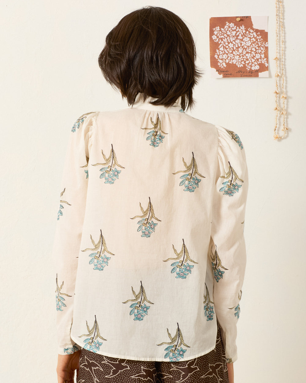 A person with short dark hair, shown from the back, wears the Artemis Sky Mist Flower Shirt—a cream-colored, long-sleeved cotton voile top with a blue floral motif. A brown artwork and beaded necklace hang on the wall.