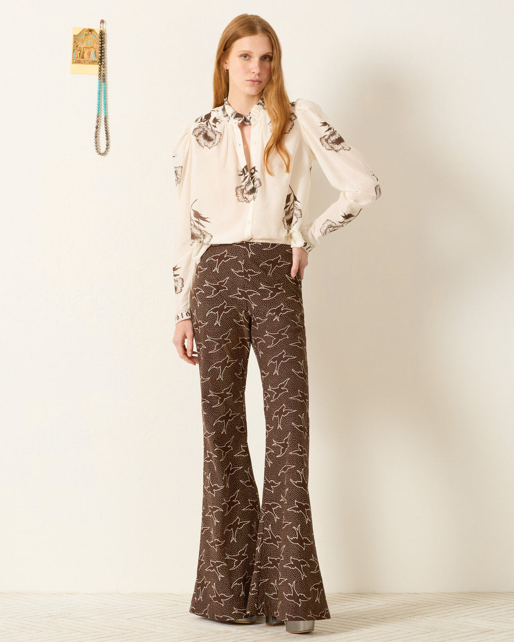 A woman stands against a plain wall wearing the Artemis Obsidian Poppy Shirt with puff sleeves and high-waisted, wide-leg brown pants featuring a white bird print.
