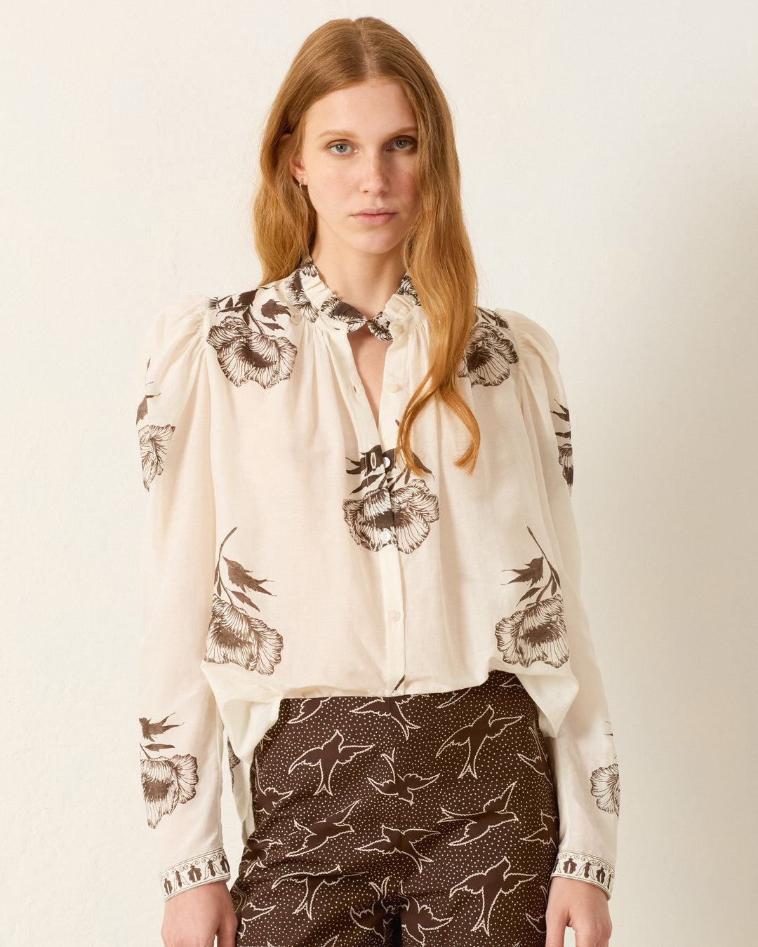 A woman with long light brown hair wears the Artemis Obsidian Poppy Shirt, a hand-block-printed blouse with dark floral prints, paired with matching pants, standing against a plain light-colored wall.