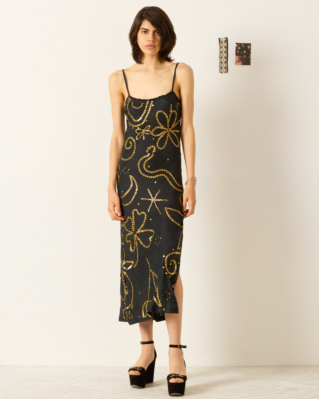 A woman poses by a white wall in the Athens Jet Coin Silk Dress, a black midi with Klimt-inspired motifs and a ruffle-trimmed neckline, paired with platform heels. Two small artworks hang on the wall beside her.