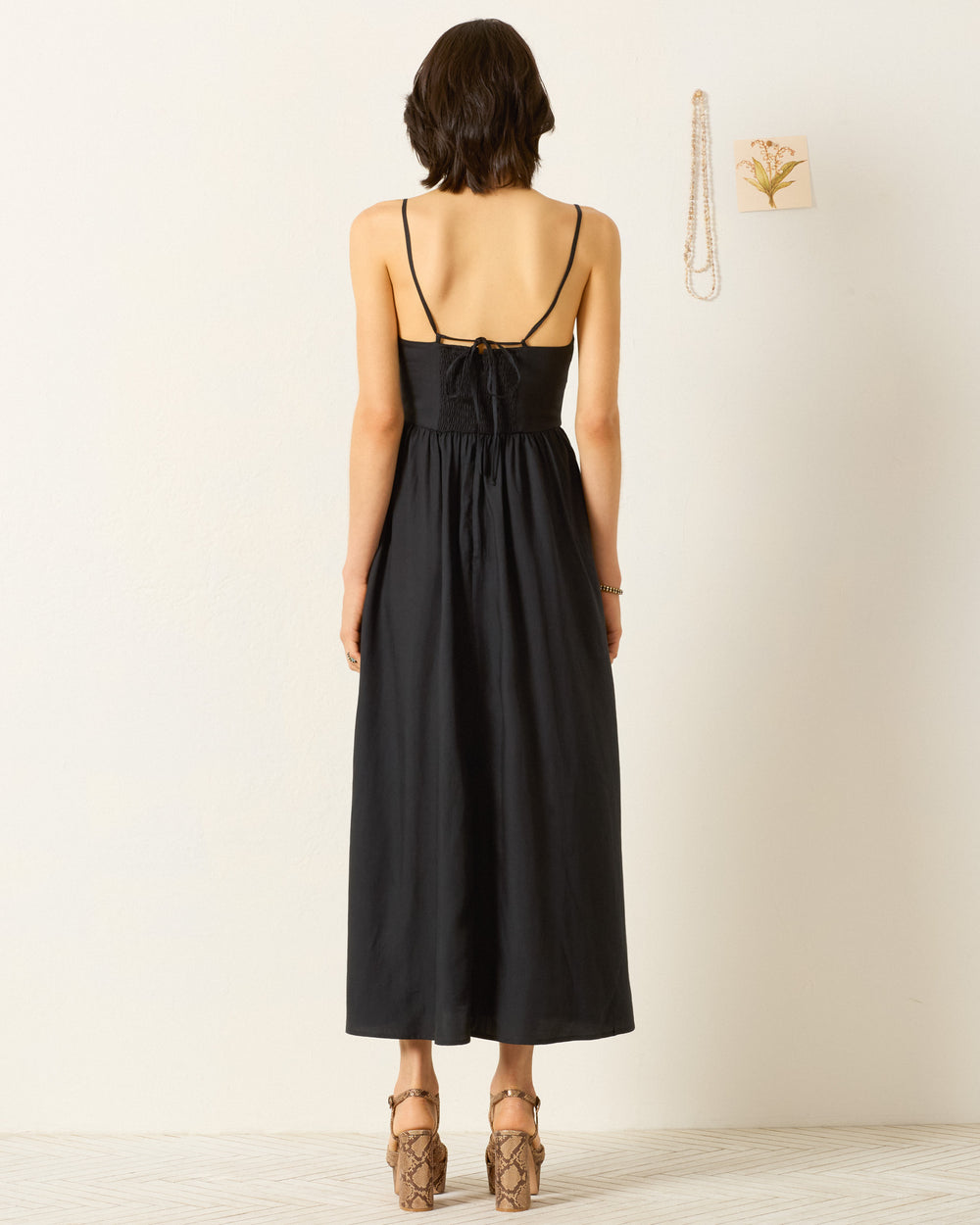 A woman stands facing away in the Autumn Abstract Bloom Dress, a Klimt-inspired black sleeveless dress with thin straps, paired with heeled sandals in a minimal room with a wall hanging and necklace.
