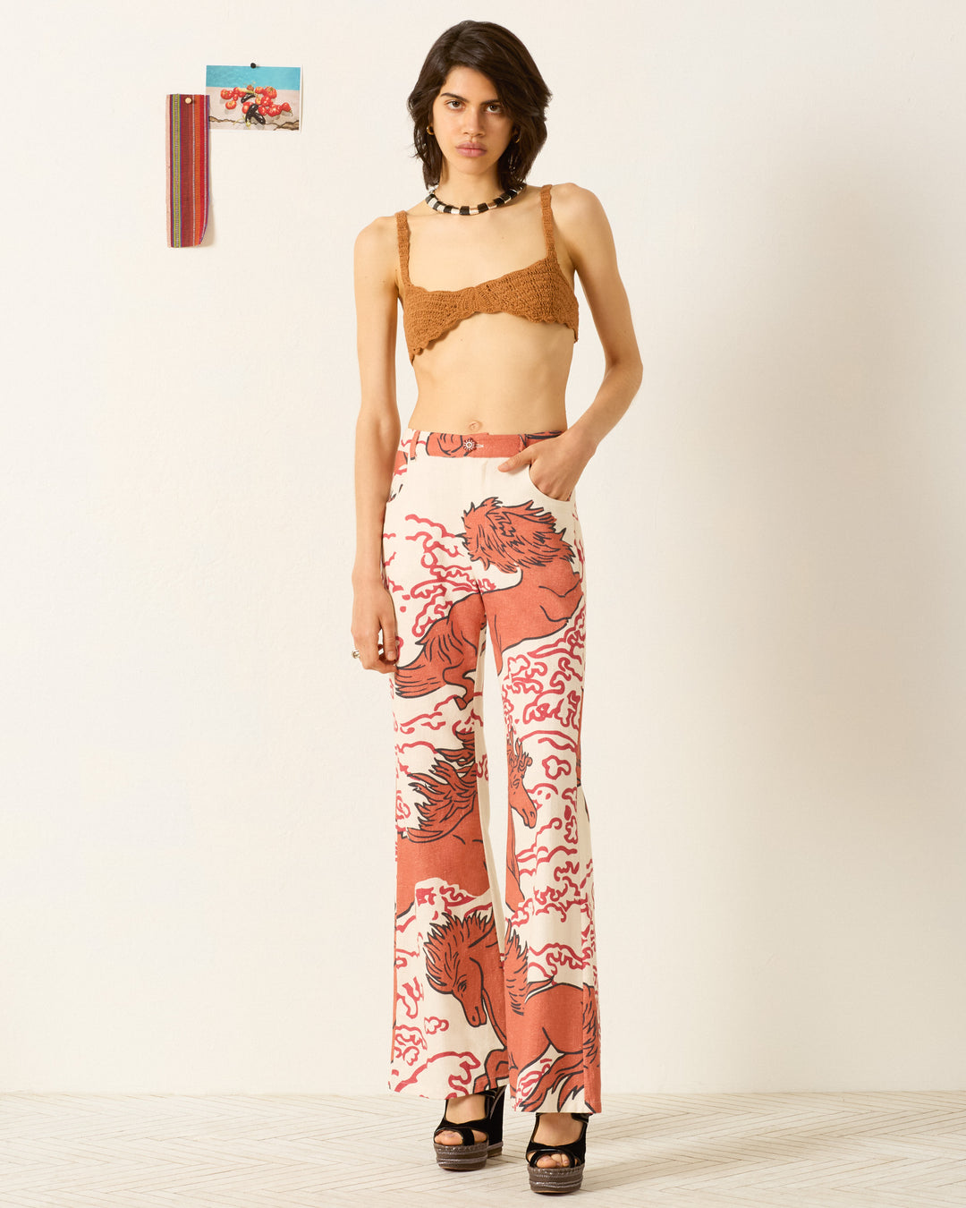 A person wears Bunny Rouge Pegasus Jean—high-waisted, wide-leg khadi cotton pants with a red and beige horse print—paired with a brown bralette and black platform sandals, standing by a plain wall with small artwork.
