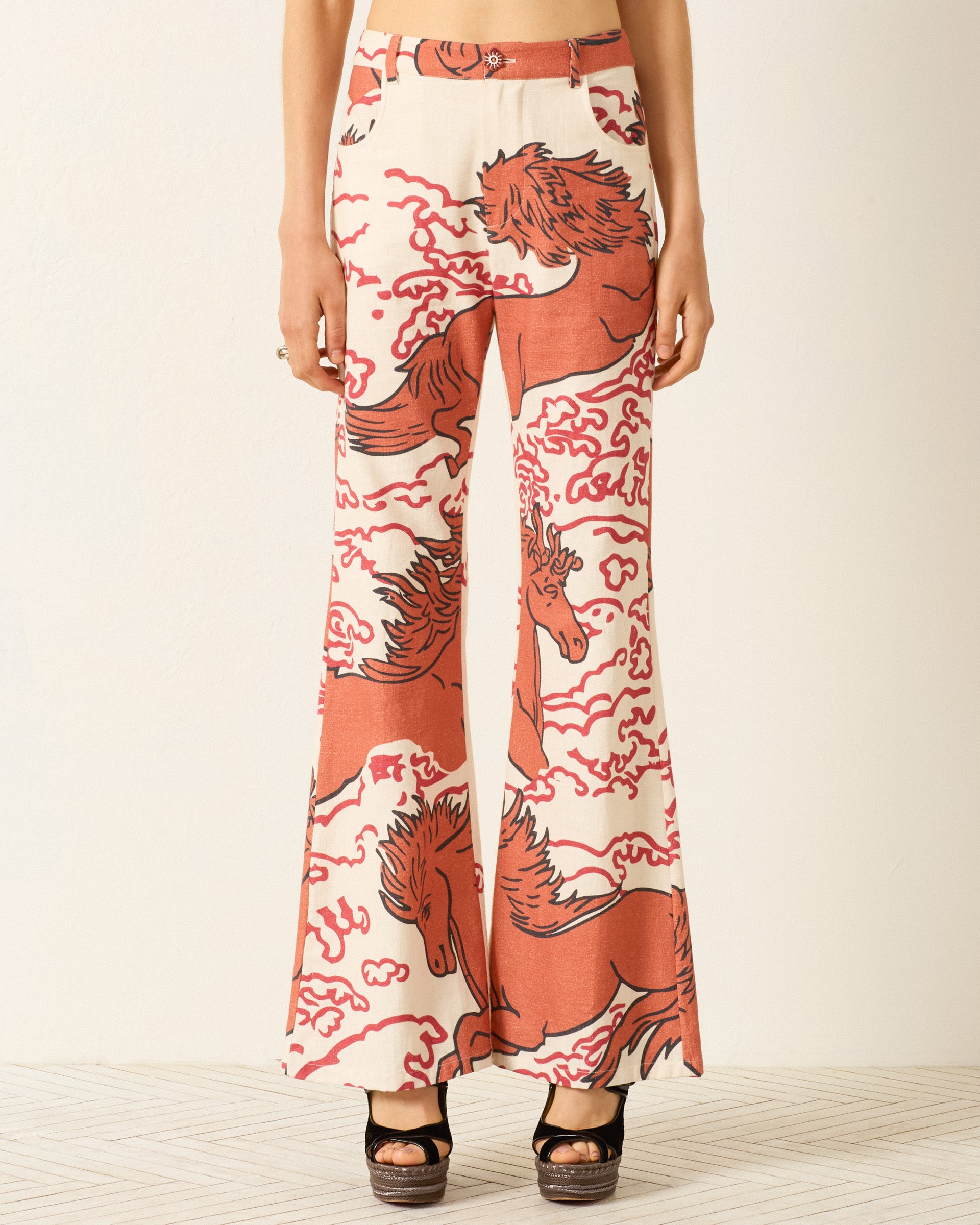 Person wearing Bunny Rouge Pegasus Jean—cream Khadi cotton flared pants with bold red horse and cloud motifs, jean-cut details, and a handmade button. Styled with black platform sandals; torso and feet visible, neutral background.