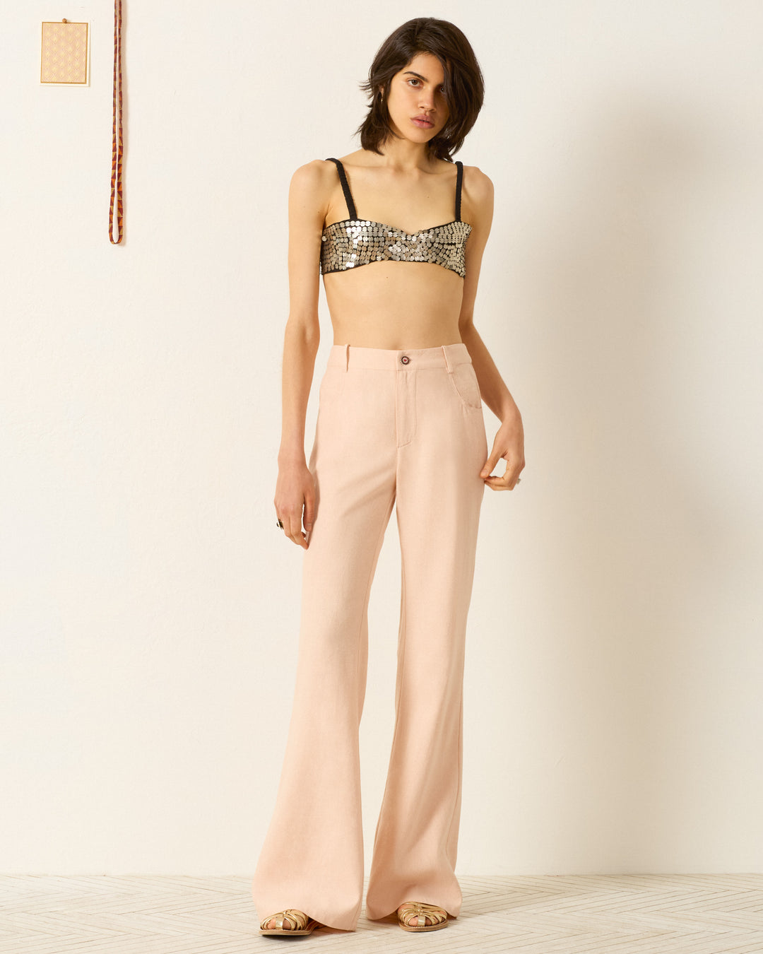 A person indoors wears a silver sequin crop top, Bunny Pink Blush Jean high-waisted light pink flared pants, and sandals against a plain background.