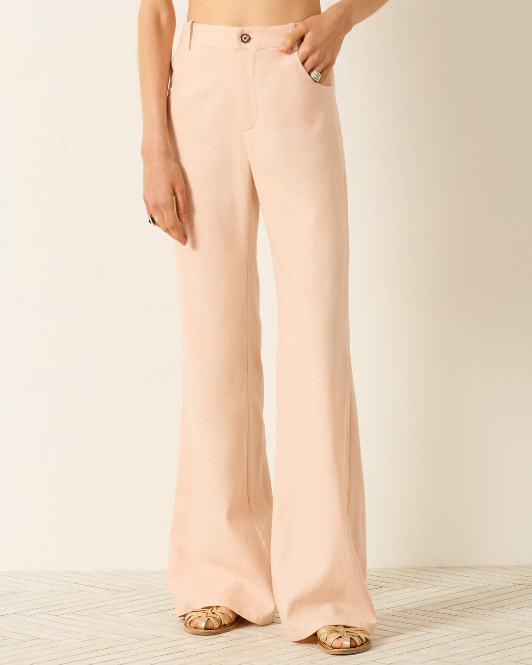 Person wearing Bunny Pink Blush Jean, a high-waisted, flared leg pant in light peach, paired with beige open-toe platform shoes, standing against a plain light background.