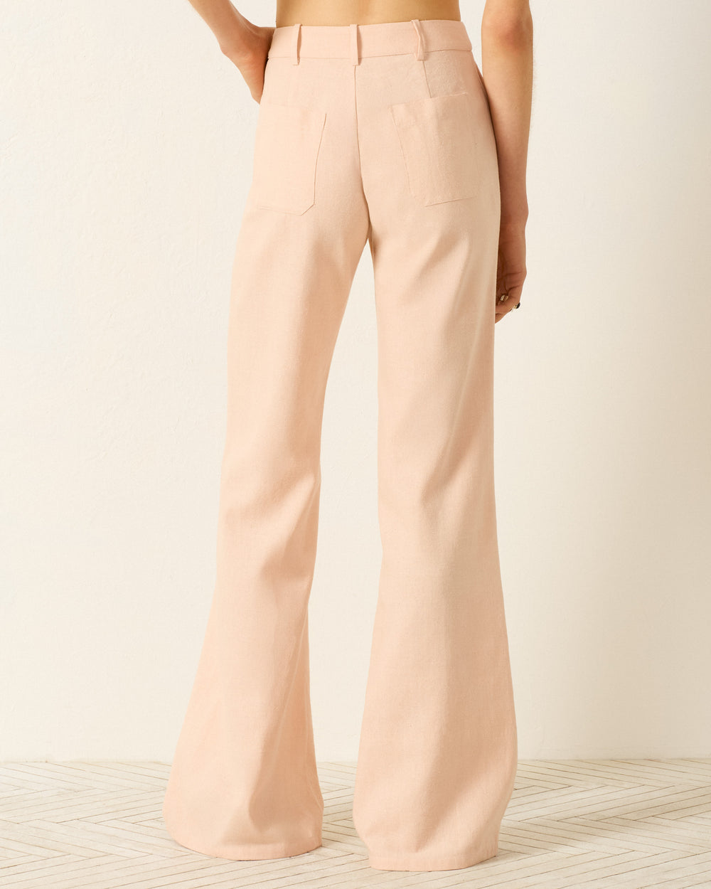 Model wearing Bunny Pink Blush Jean, a high-waisted, wide-leg, light pink flared pant, photographed from the back against a neutral background.