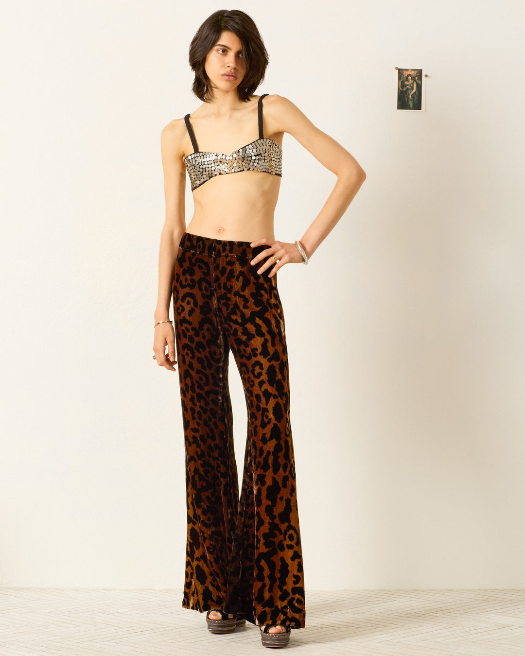 Indoors, a person wears a sequined bralette, platform sandals, and the Farah Leopard Velvet Pant—high-waisted velvet bell-bottoms with a leopard print. A small photo is taped to the wall behind them.