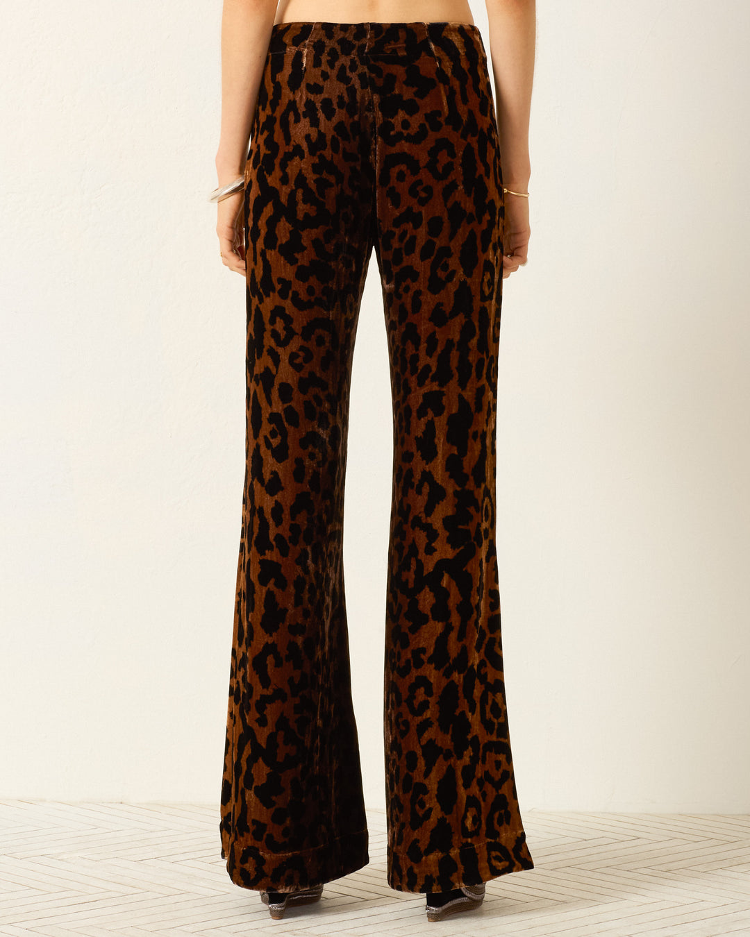A person stands with their back to the camera, wearing the Farah Leopard Velvet Pant—high-waisted pants in bold brown and black leopard-print velvet, featuring a dramatic bell-bottom flare.