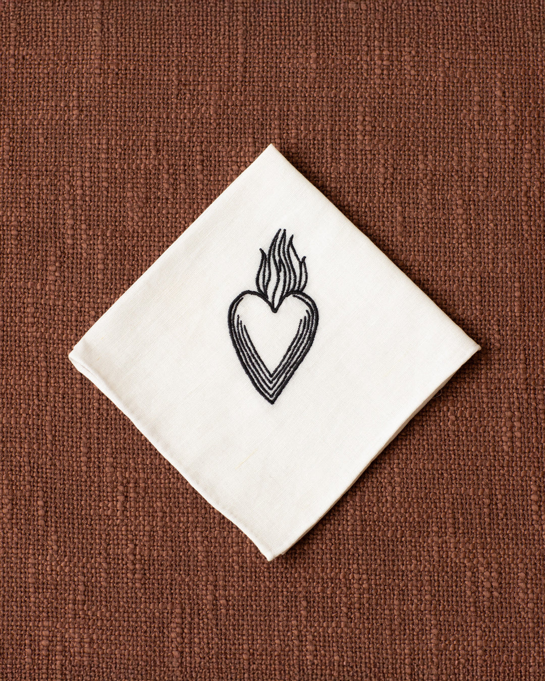 The Cai Burning Heart Handkerchief, made of white linen-cotton with a black embroidered heart and flames, is folded into a square and displayed on a textured brown surface—a distinctive token of love.