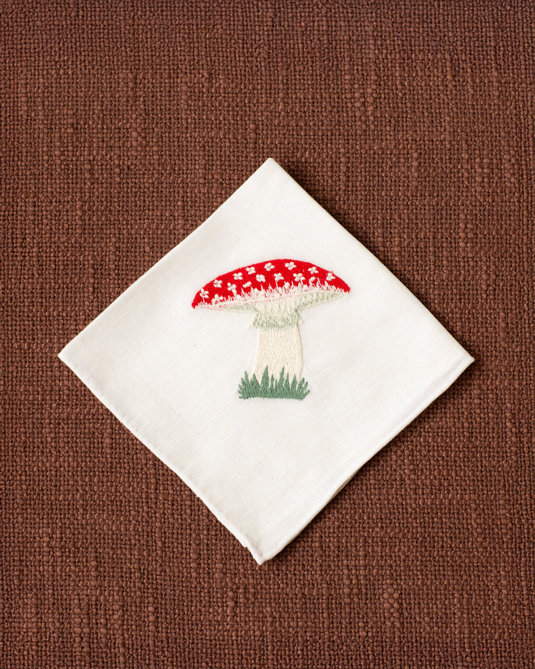 The Cai Mushroom Handkerchief, made of white linen-cotton and embroidered with a red and white fly agaric toadstool, rests on a textured brown surface.