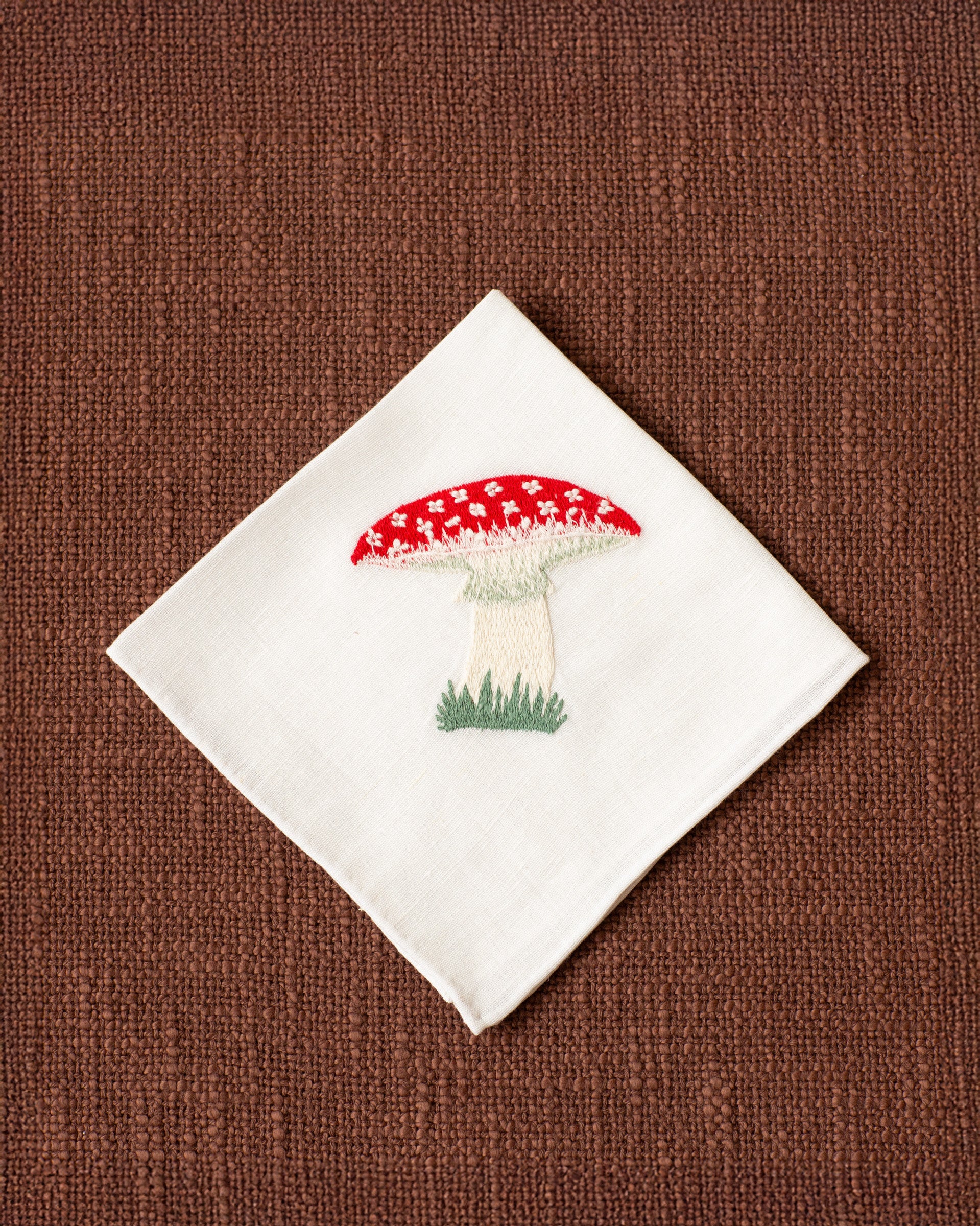The Cai Mushroom Handkerchief, made of white linen-cotton and embroidered with a red and white fly agaric toadstool, rests on a textured brown surface.