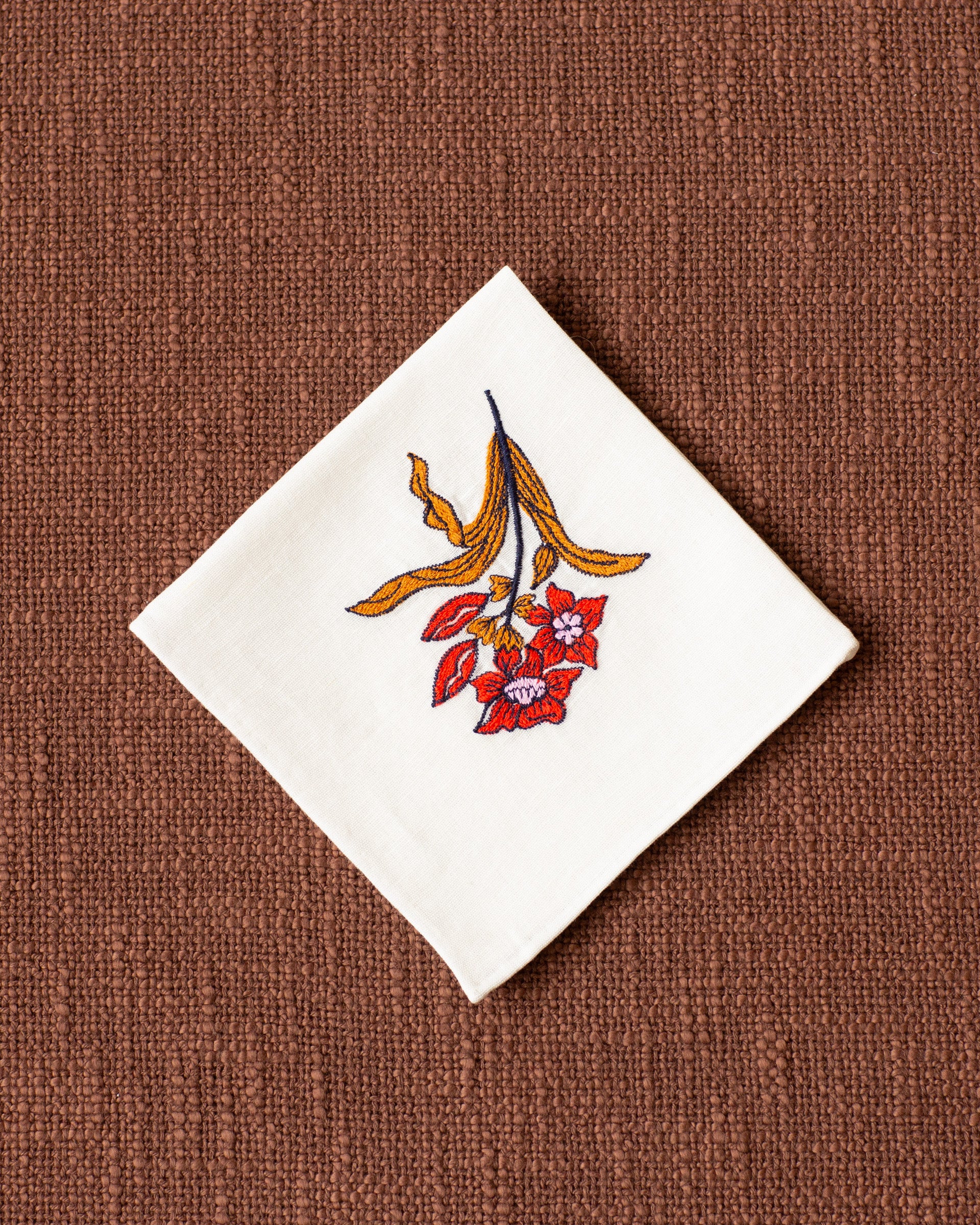 The Cai Little Flower Handkerchief, made from white linen-cotton and adorned with red and yellow floral embroidery, rests on a textured brown surface—an elegant gift for good luck.