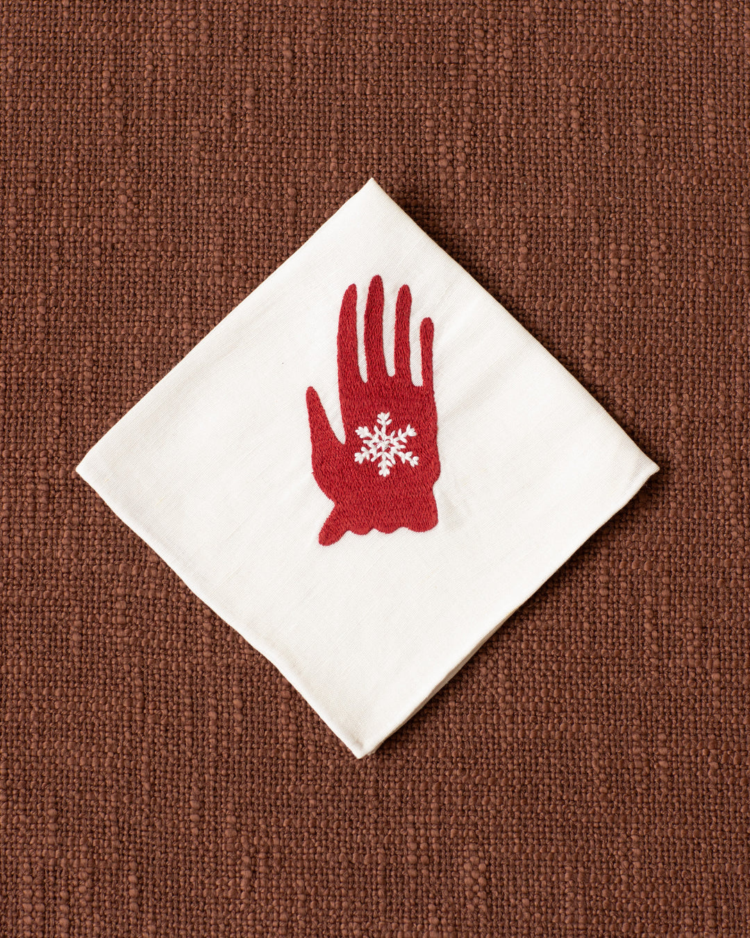 The Cai Handshake Handkerchief, made from white linen-cotton, features a red hand and snowflake design and is displayed diagonally on a textured brown surface.