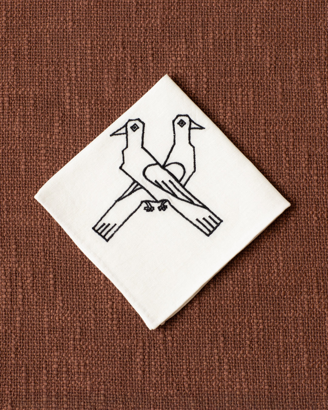 The Cai Lovebird Handkerchief, crafted from linen-cotton and adorned with a black outline of two lovebirds facing opposite directions, rests on a textured brown surface—making it a unique gift of love.
