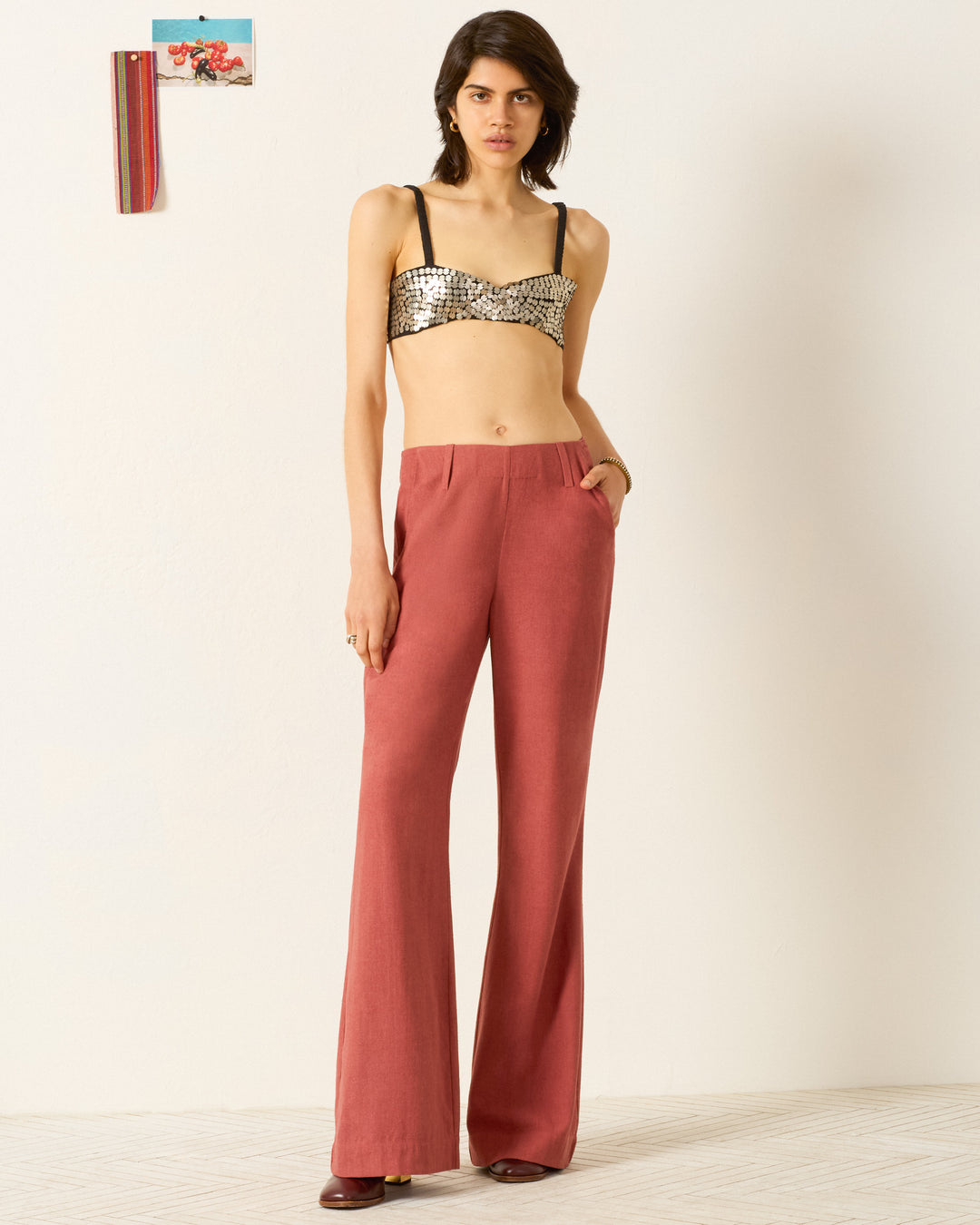 A woman stands against a white wall wearing a metallic crop top and high-waisted Charlie Red Ochre Silk Pant with flared legs, her left hand tucked in her pocket.