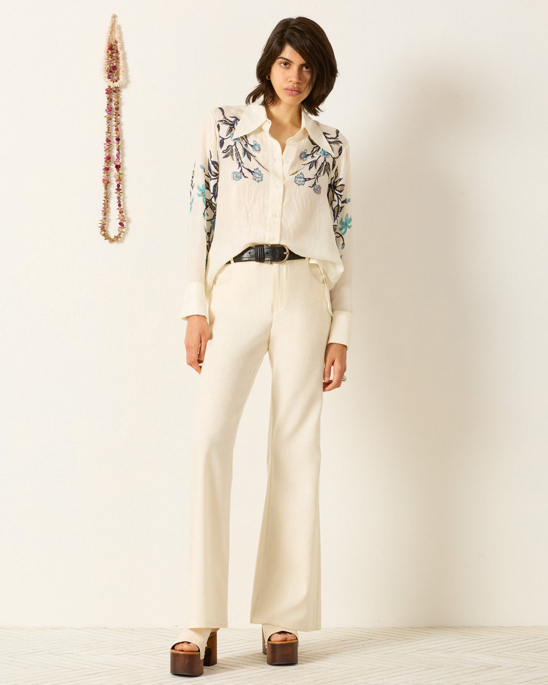 A person stands indoors wearing the Dana Misted Lily Silk Shirt, cream-colored wide-leg pants, a black belt, and platform sandals; a beaded necklace hangs on the wall behind them.