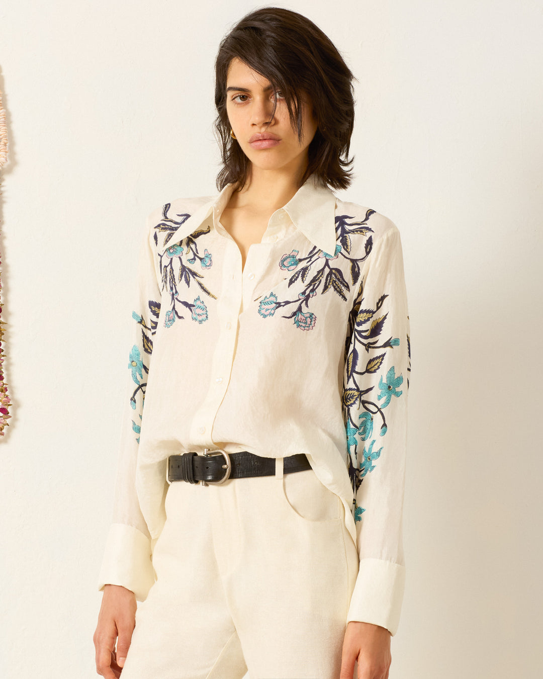 A person wearing the Dana Misted Lily Silk Shirt—cream-colored, hand-loomed silk with blue floral embroidery—paired with cream pants and a black belt, standing against a plain light background.