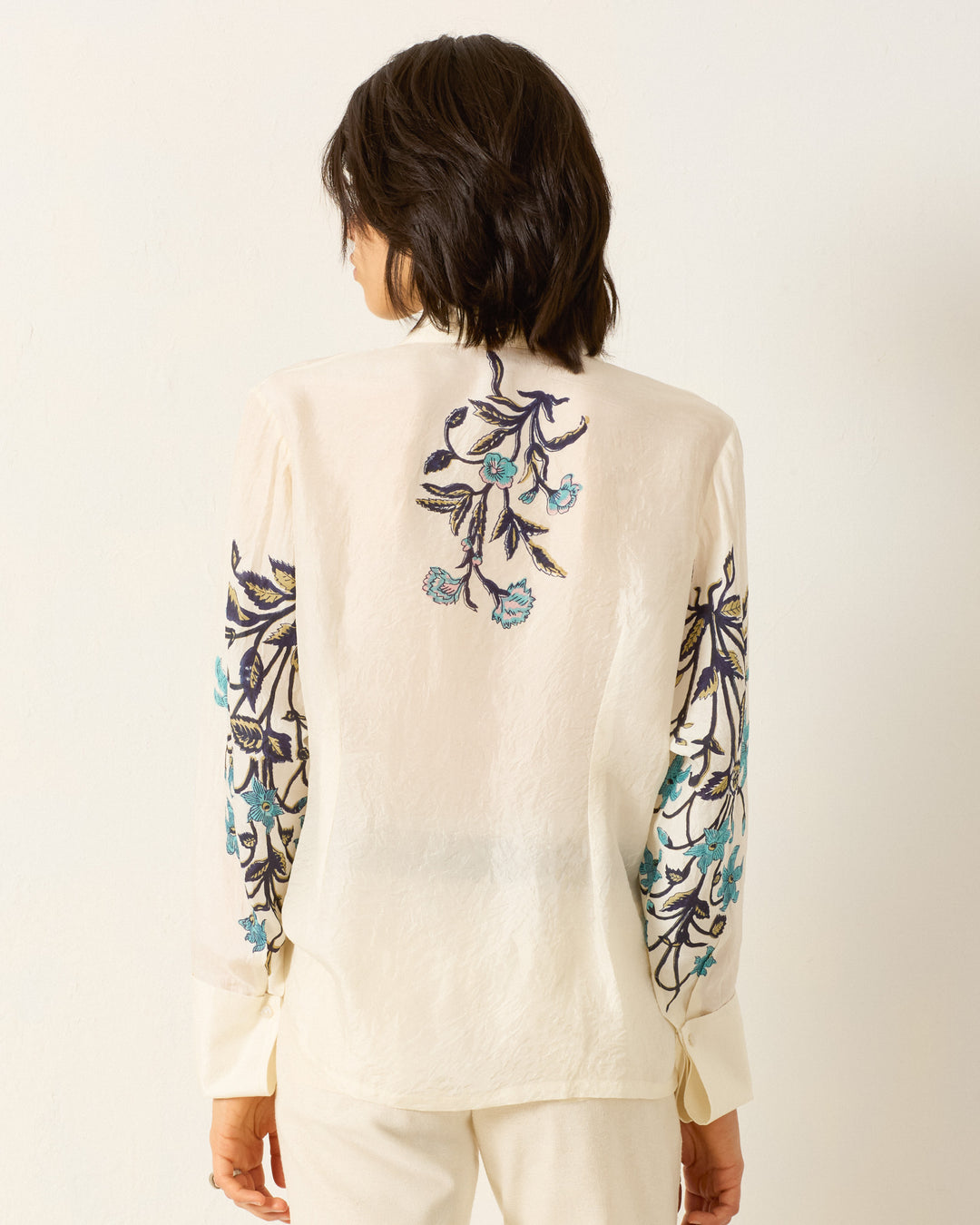 A person is seen from behind in the Dana Misted Lily Silk Shirt, featuring a cream color with blue and black floral embroidery on the back and sleeves, set against a plain light background.