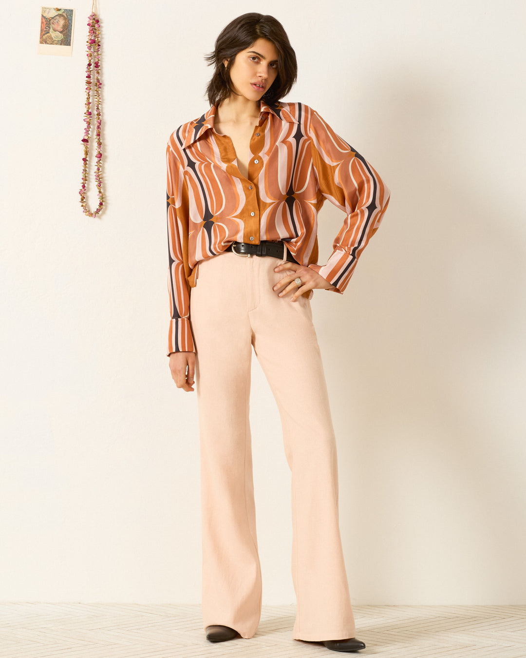 A person stands against a white wall wearing the Dana Mesa Verde Silk Shirt with a retro print and '70s dagger collar, paired with light beige flared pants and black shoes, one hand on their hip.