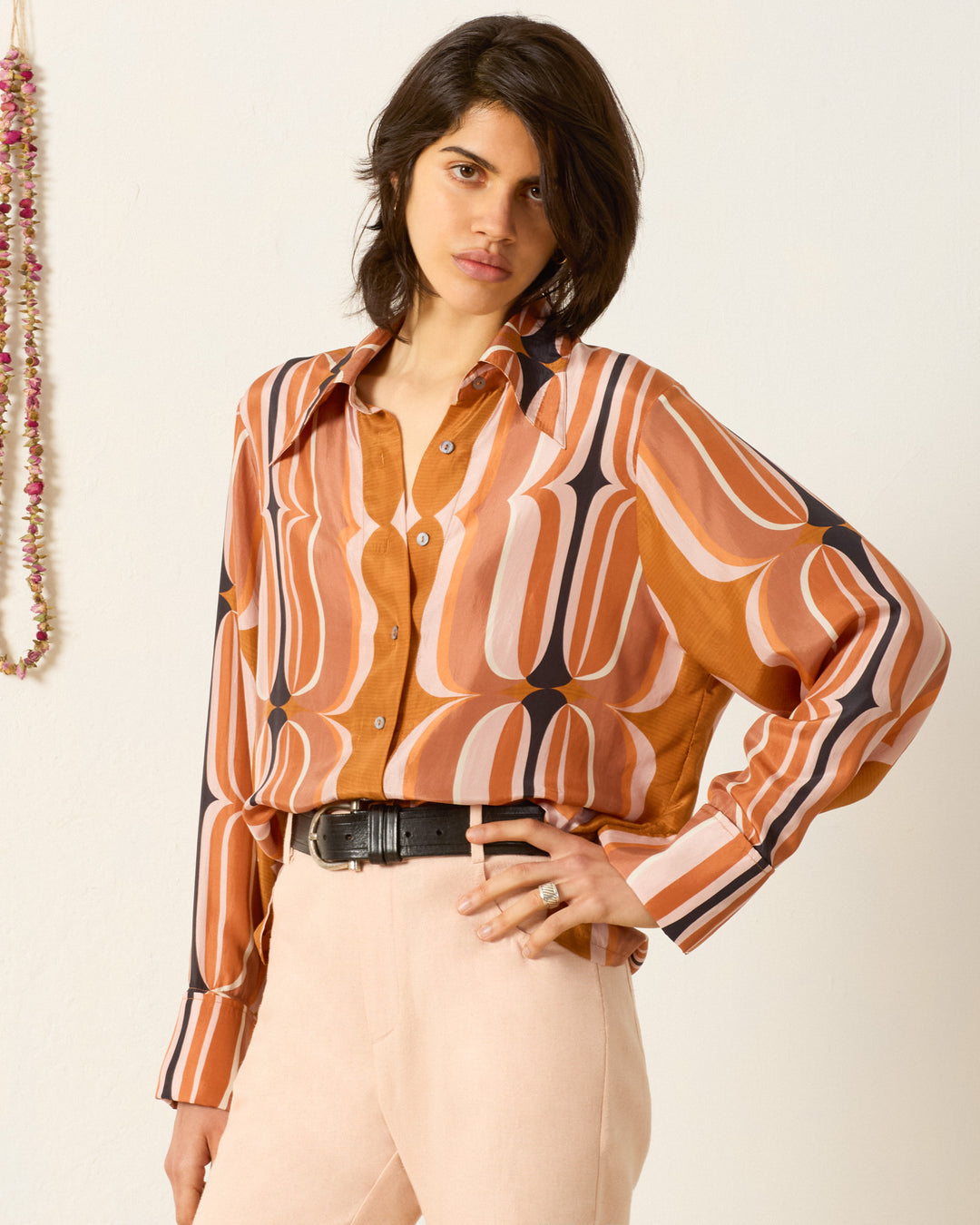 A person stands against a plain wall wearing the Dana Mesa Verde Silk Shirt, a retro-inspired, long-sleeve button-up in warm tones, paired with high-waisted, light-colored pants and a black belt.