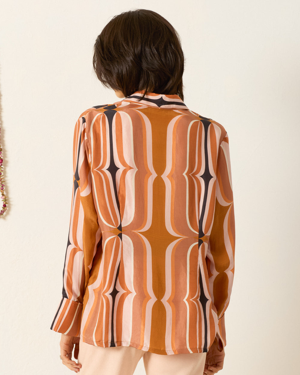 Model wearing the Dana Mesa Verde Silk Shirt—a retro-inspired long-sleeve, button-up with an orange, pink, and black geometric print and '70s dagger collar—shown from the back against a plain light wall.