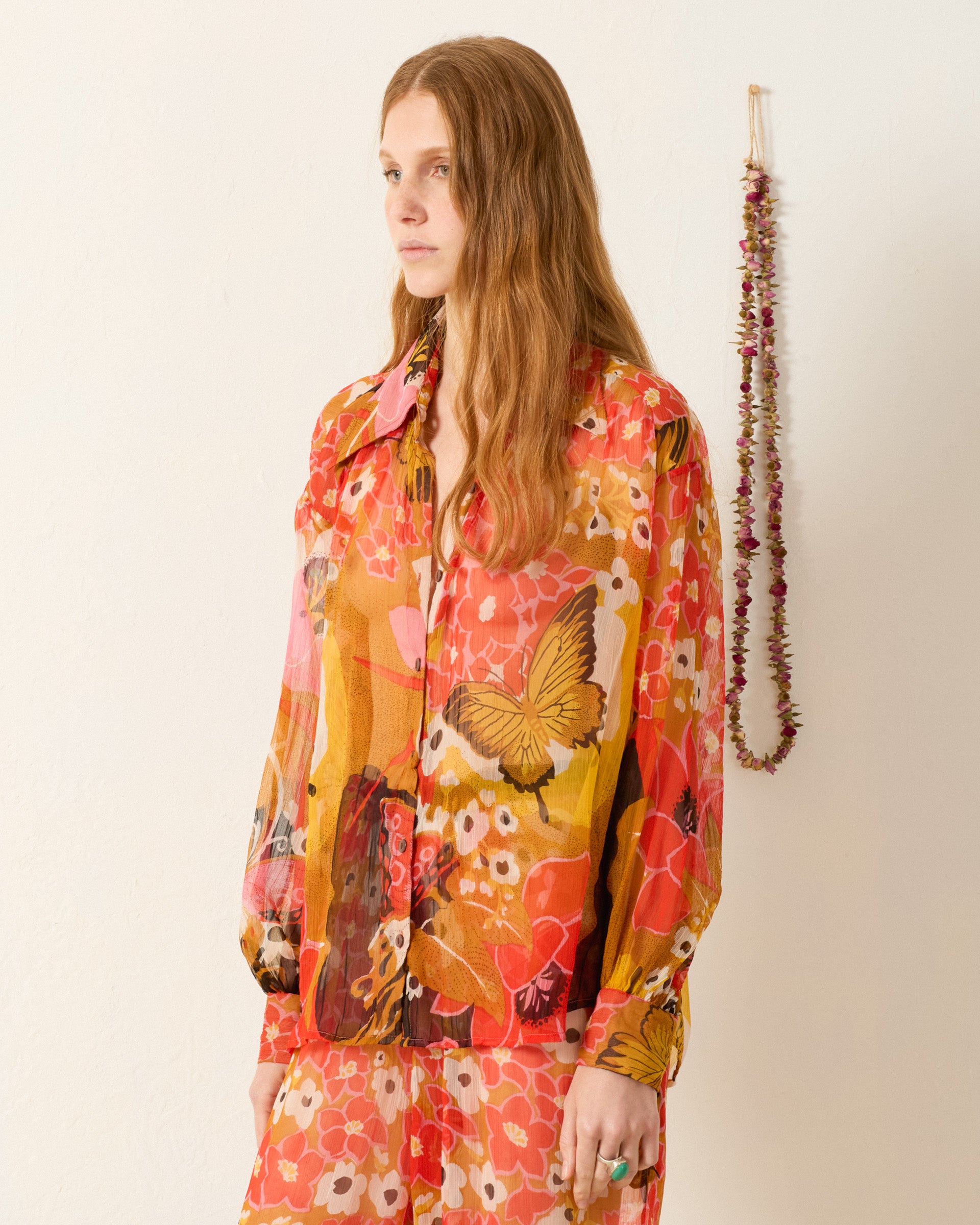 A woman with long, wavy red hair stands indoors by a hanging garland, wearing the vintage-inspired Emery Marigold Meadow Shirt featuring a sheer floral and butterfly print.