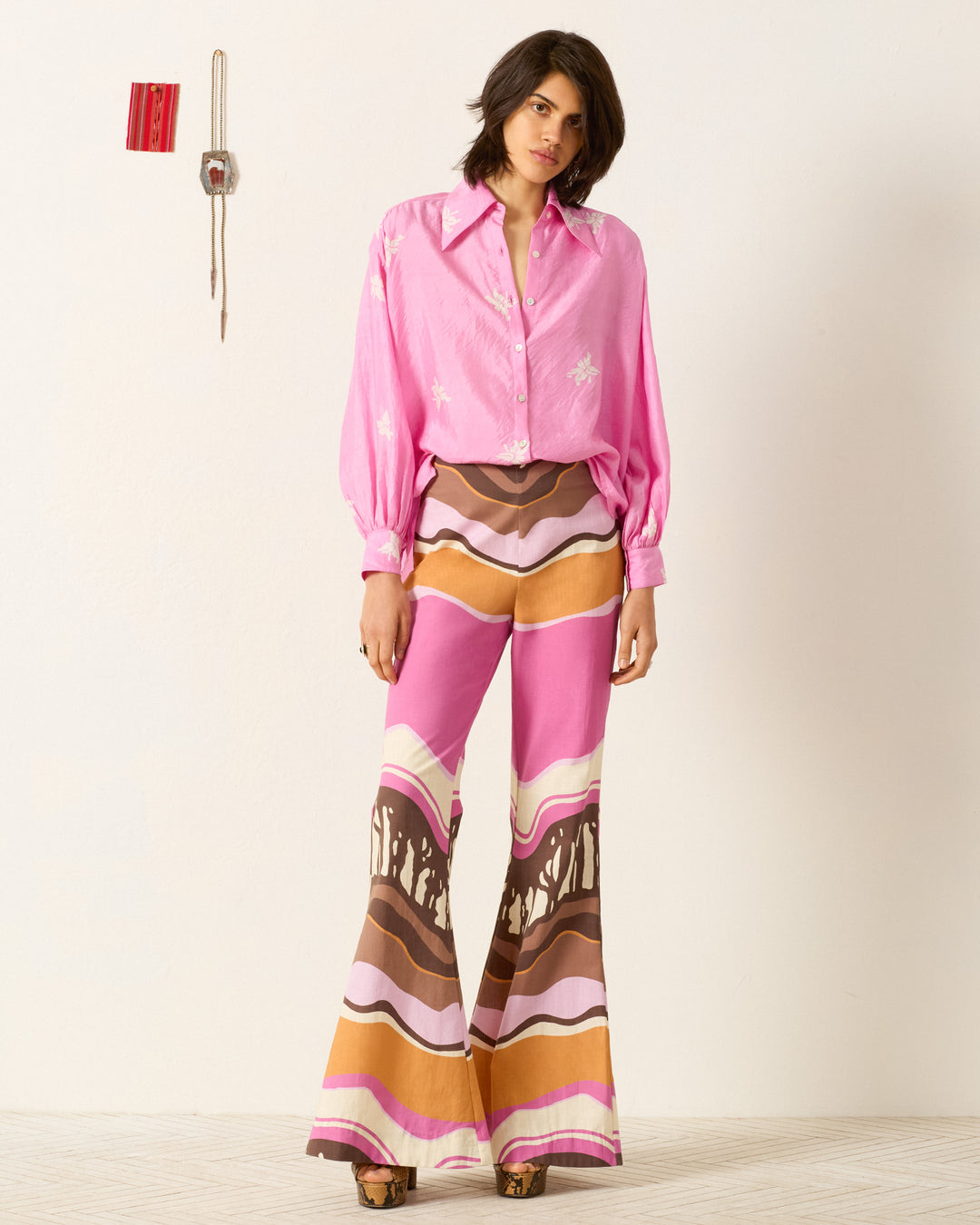 Indoors, a woman wears the Emery Pink Jasmine Shirt, paired with high-waisted flared pants featuring a colorful wavy pattern. A red wall fixture and cord are visible on the wall behind her.