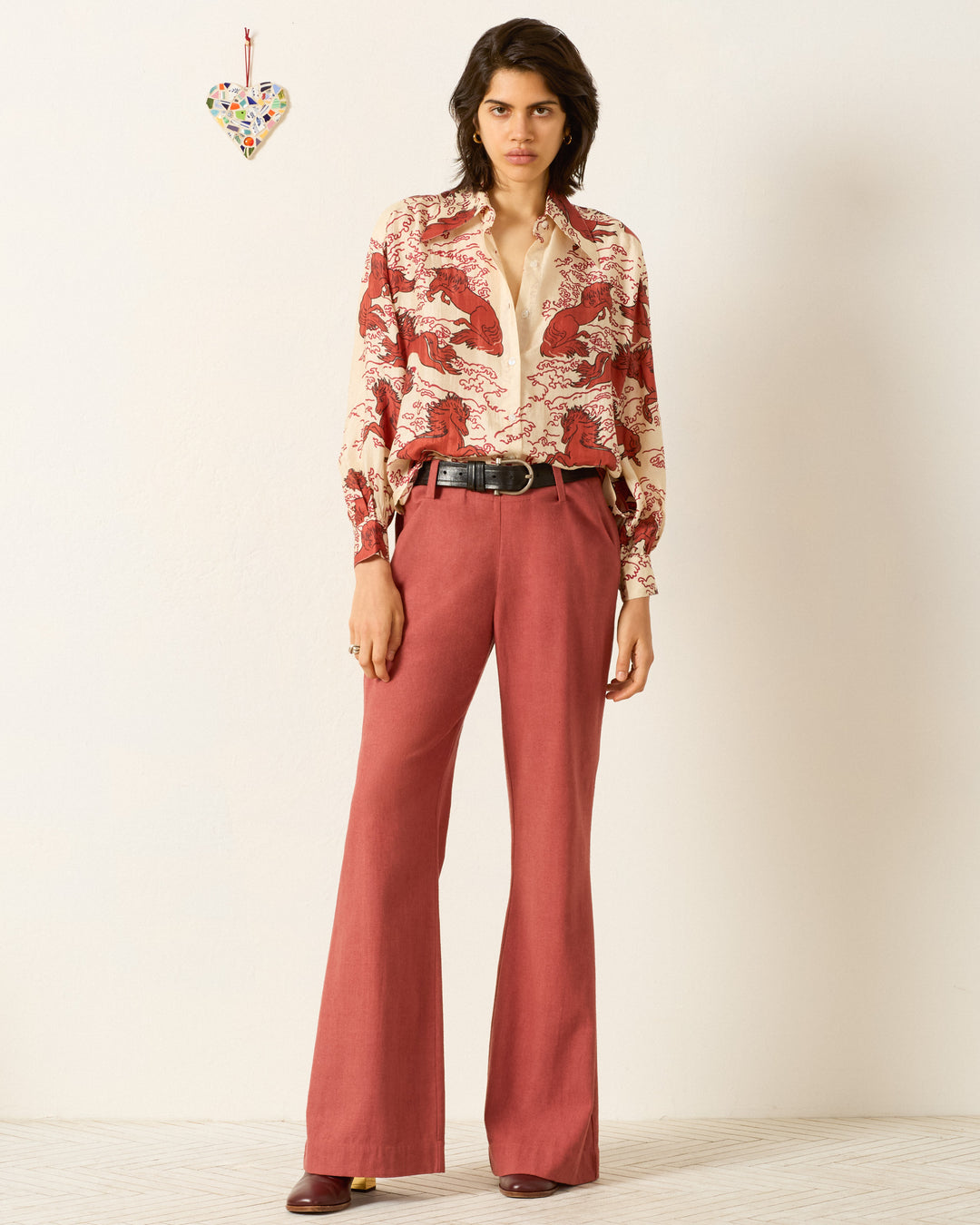 A person stands against a plain wall wearing the artisan-made Emery Rouge Pegasus Shirt, featuring a mythical horse motif, paired with high-waisted red trousers, a black belt, and red shoes. A decorative heart hangs on the wall.