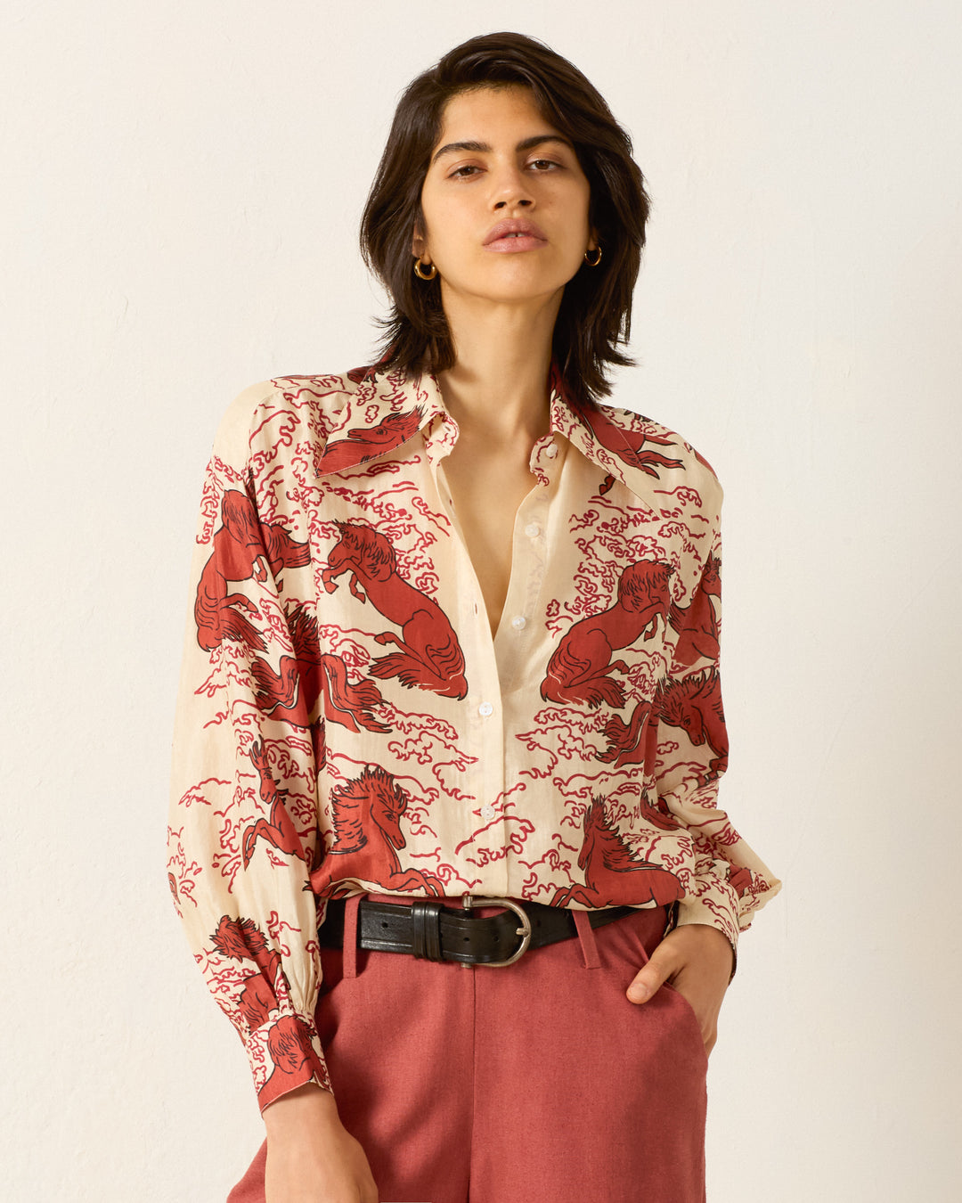 A person stands against a plain wall wearing the artisan-made Emery Rouge Pegasus Shirt, featuring a mythical horse and red illustrations, paired with pink trousers and a black belt.