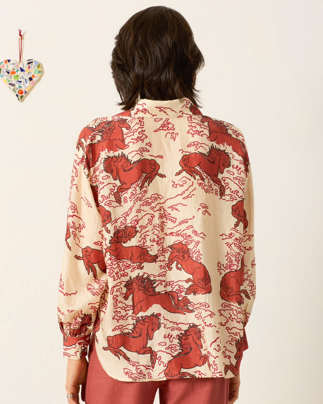 A person with dark hair, seen from behind, wears the Emery Rouge Pegasus Shirt—a hand-loomed silk top featuring a red mythical horse motif—paired with rust-colored pants. A heart-shaped decoration hangs on the left wall.