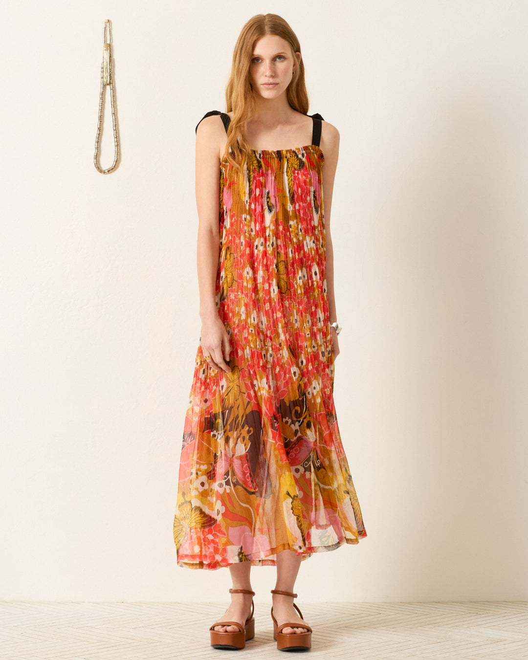 A woman stands against a plain wall wearing the Emrys Marigold Meadow Dress with black shoulder straps and brown platform sandals. A rope hangs on the wall to her left.