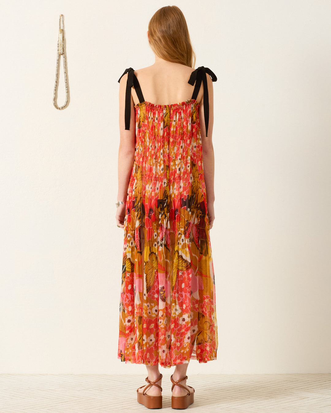 A woman with long hair faces away, wearing the Emrys Marigold Meadow Dress—a colorful floral maxi with ruched tiers and black shoulder ties—paired with platform sandals in a minimalist room.