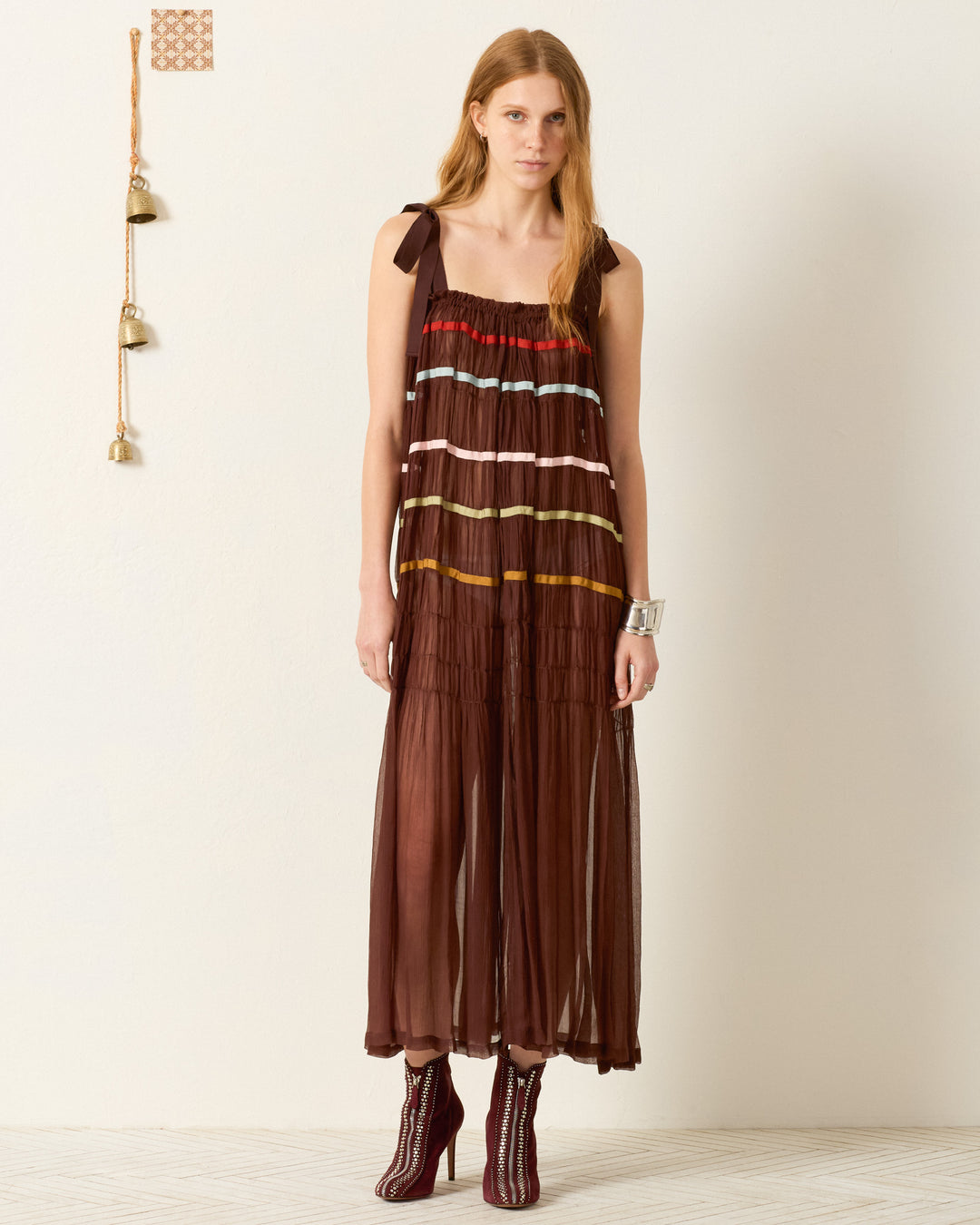 A woman stands indoors wearing the Emrys Clove Aurora Dress, a sleeveless sheer brown cupro-chiffon maxi with colorful stripes. She pairs it with ankle boots and a silver cuff. Three bells hang on the wall beside her.