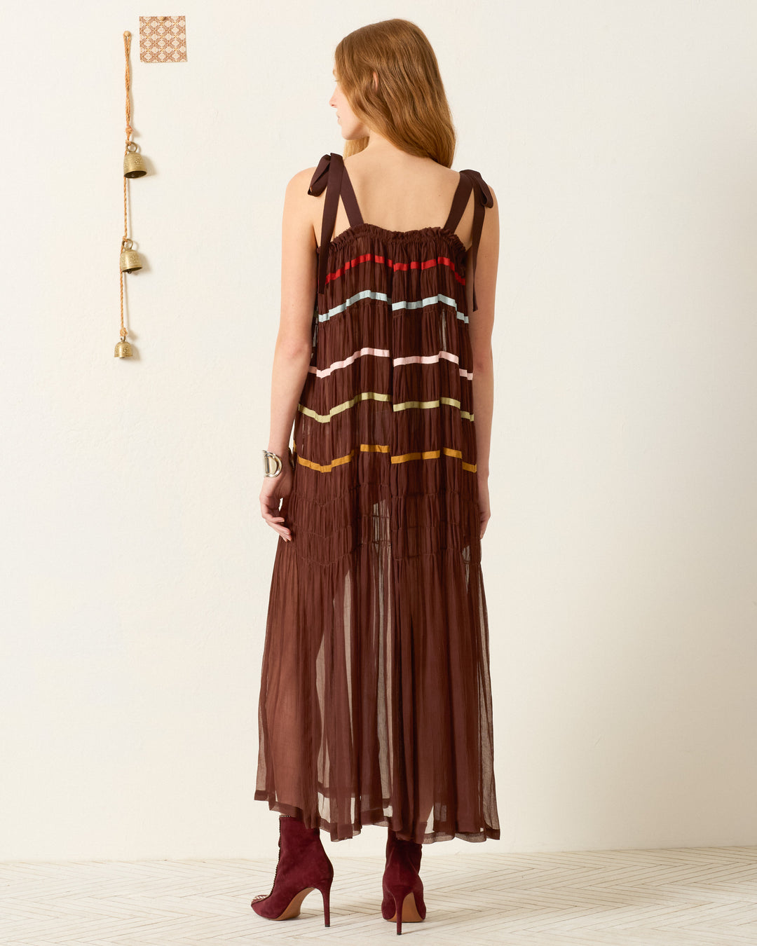A woman stands facing away, wearing the Emrys Clove Aurora Dress—a sleeveless cupro-chiffon maxi with colorful horizontal stripes and tie straps—paired with maroon heeled boots.