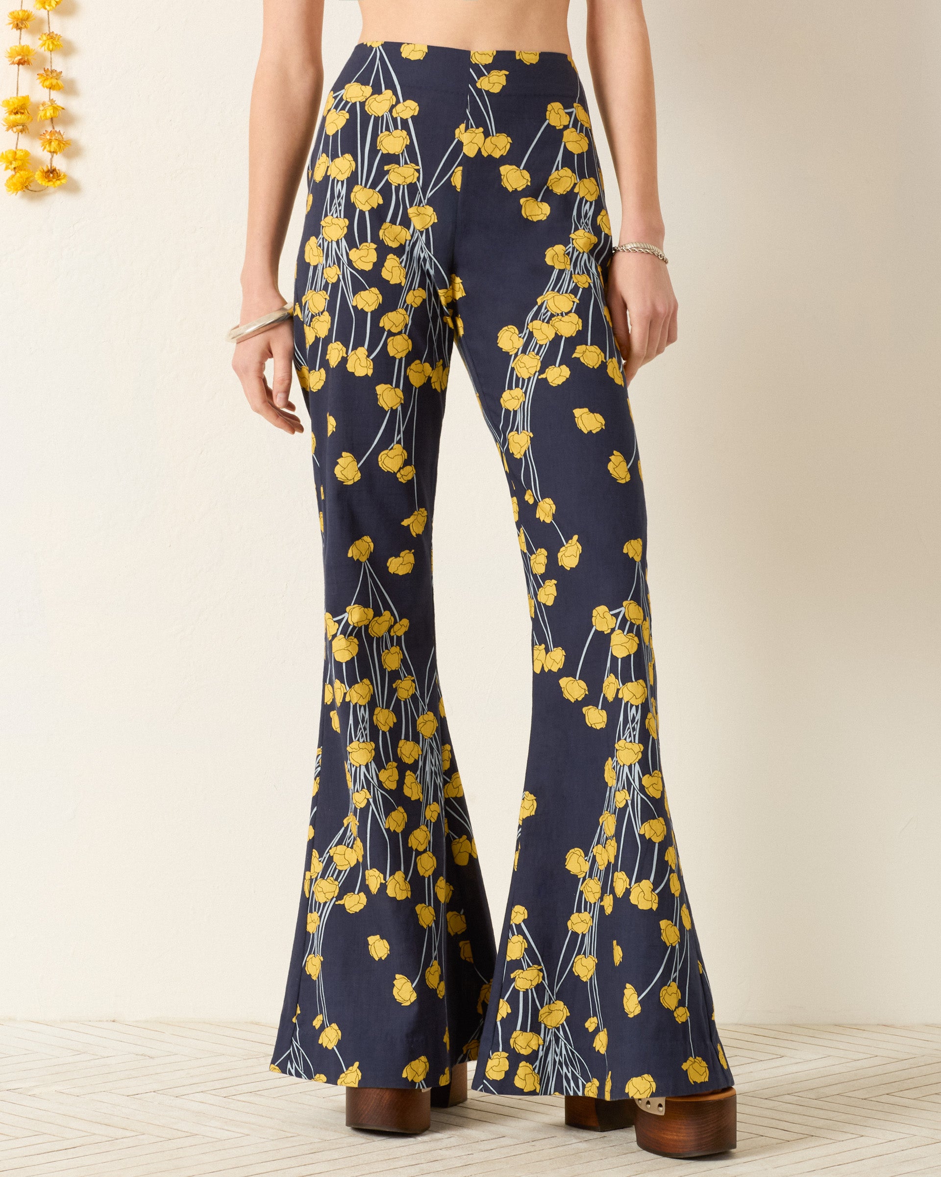 A woman wears the Farah Buttercup Bud Pant—high-waisted, wide-leg pants with a navy blue base and buttercup print—paired with platform shoes.