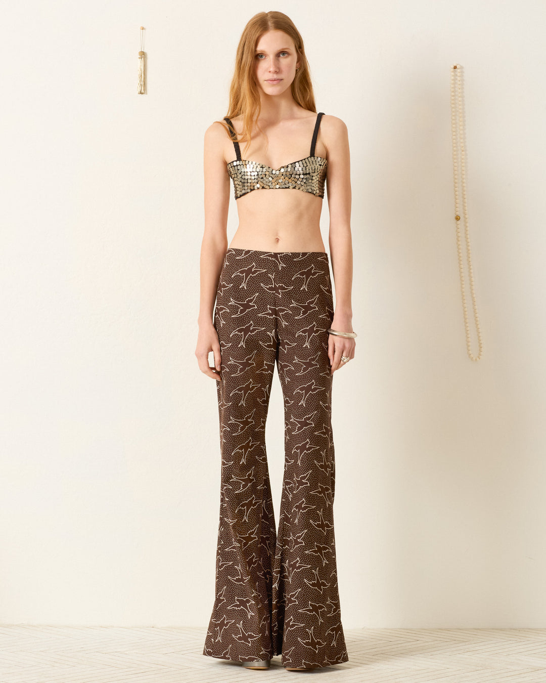 A woman stands indoors facing the camera in the Farah Clove Swallow Pant, featuring a high waist and swallow motif. Her look is completed by a sequined bra top and dramatic bell-bottom flare against a plain wall.