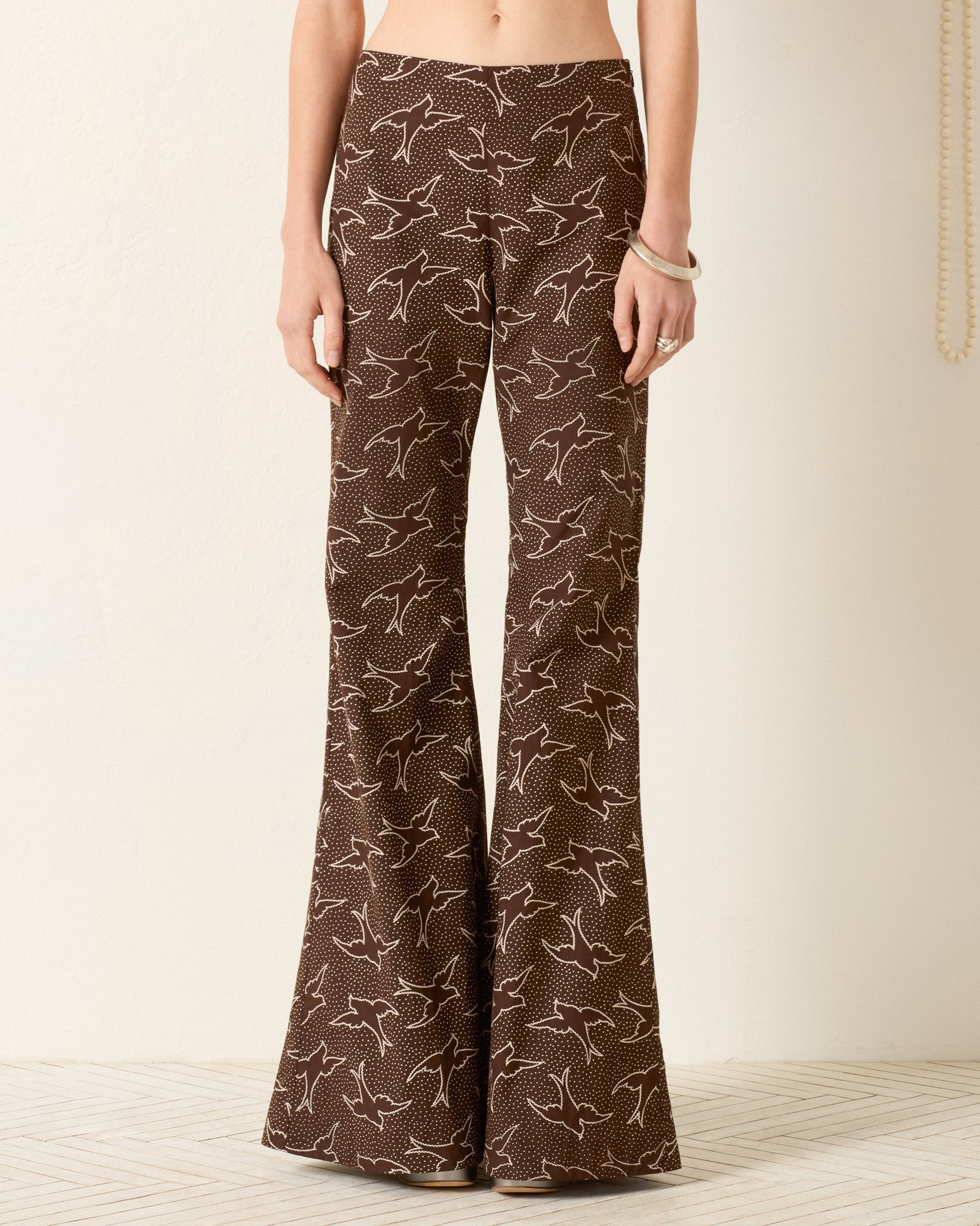 Wearing the Farah Clove Swallow Pant—high-waisted, brown, flared pants with a white swallow motif—while standing against a light-colored wall.