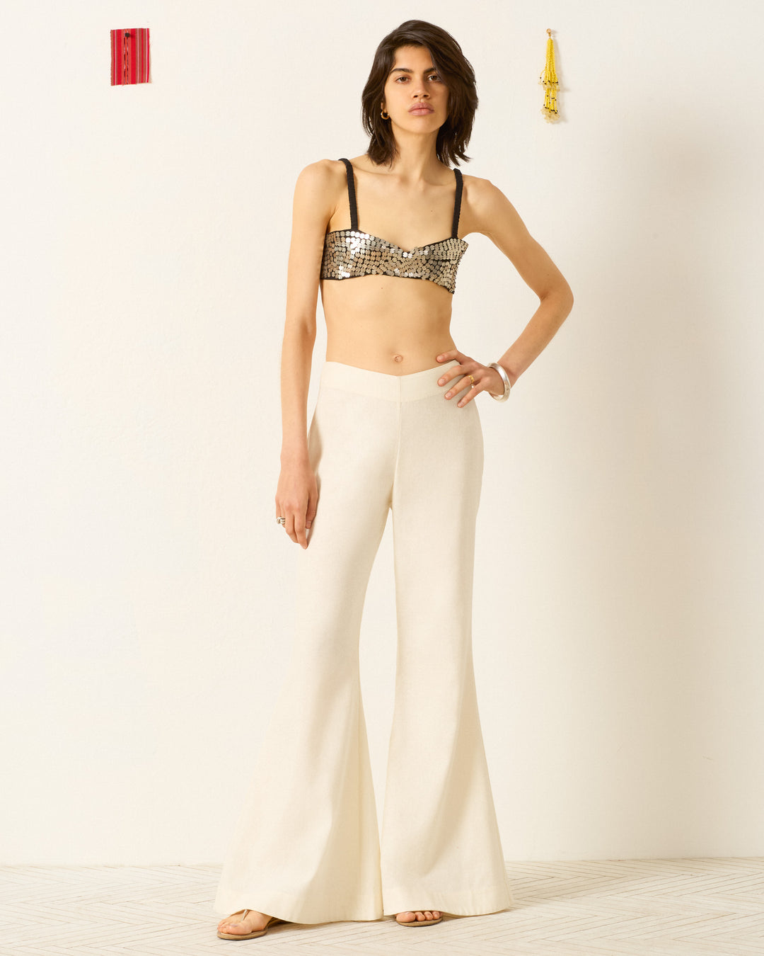 A woman with short brown hair and a neutral expression stands against a white wall, wearing the Farah Alabaster Silk Pant styled with a sequined crop top. The pants feature a high waist and subtle bell-bottom flare.