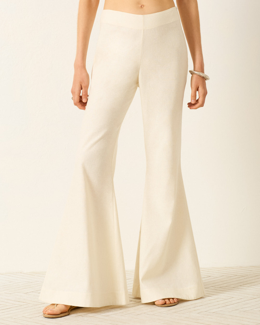 Wearing Farah Alabaster Silk Pant—high-waisted with a subtle bell-bottom flare—and sandals, this person stands on a light patterned floor against a neutral background.