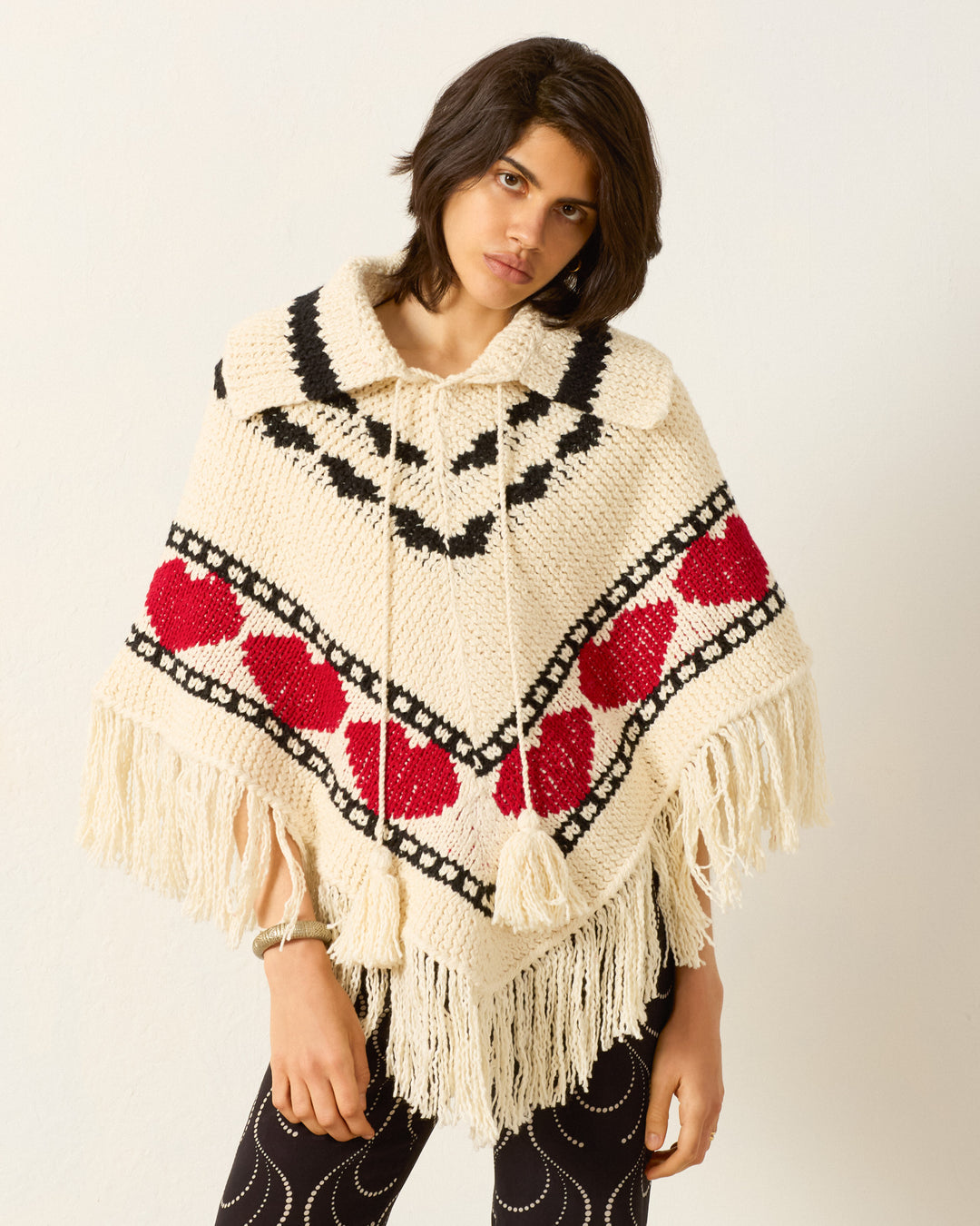 A person models the Faune Heart Knit Poncho, featuring cream fabric with red heart motifs, black patterns, and fringe, set against a plain white background.