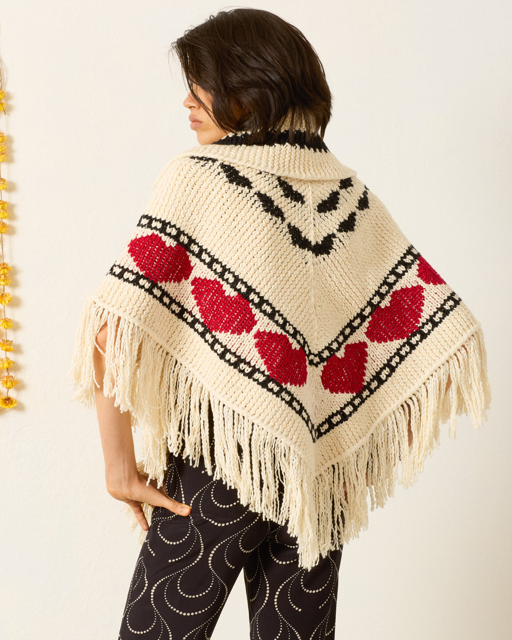A person stands with their back to the camera, showcasing cozy layering in the Faune Heart Knit Poncho—cream-colored with red heart motifs, black geometric designs, and fringe.
